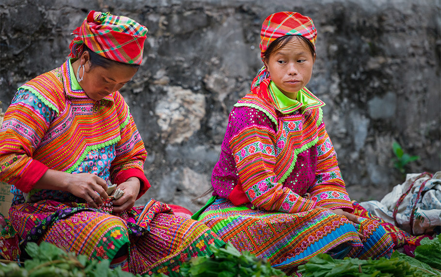 Two H'mong women sitting together wearing colorful, intricately embroidered traditional ethnic costumes in Ha Giang.