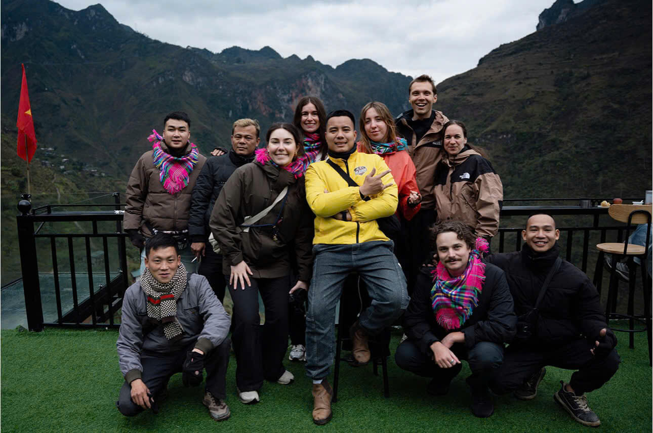 A small group of international travelers and their local Easy Rider guides posing together on a terrace overlooking the Ha Giang mountains. The group is dressed for trekking and motorbike travel, showing the intimate scale of a 2026 Ha Giang Loop tour.