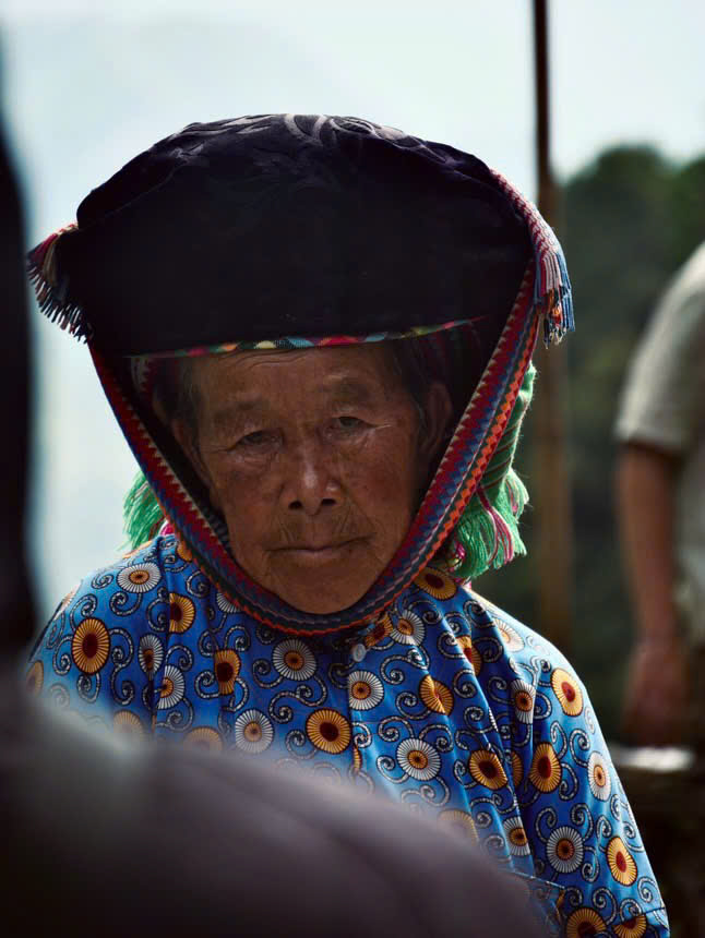 A close-up portrait of an elderly ethnic woman wearing a traditional dark headdress and a blue patterned tunic in Ha Giang.