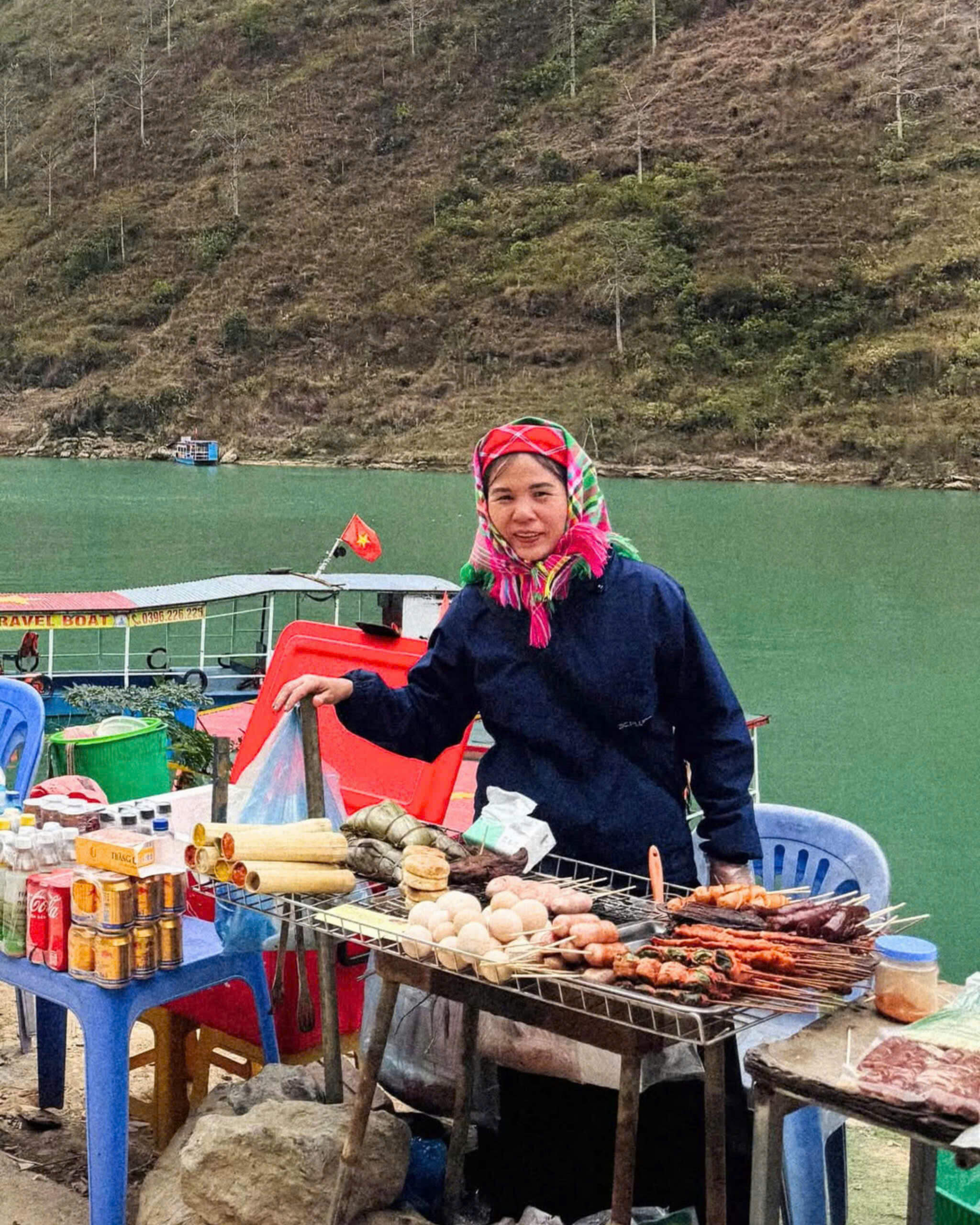 A local woman in traditional attire selling grilled street food by the Nho Que River, showcasing the culinary culture of ethnic groups.