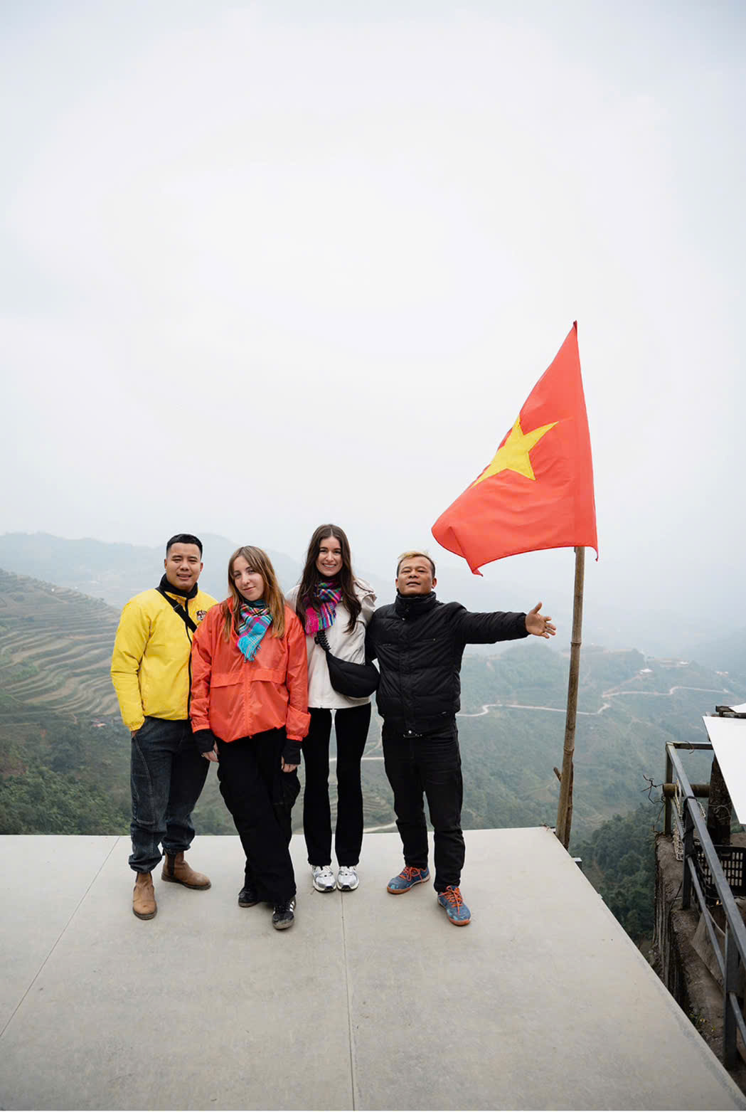 A small group of four travelers and local guides posing on a mountain overlook during a Ha Giang Loop tour in Vietnam. They stand next to a waving Vietnamese flag with a backdrop of misty mountains and terraced rice fields.