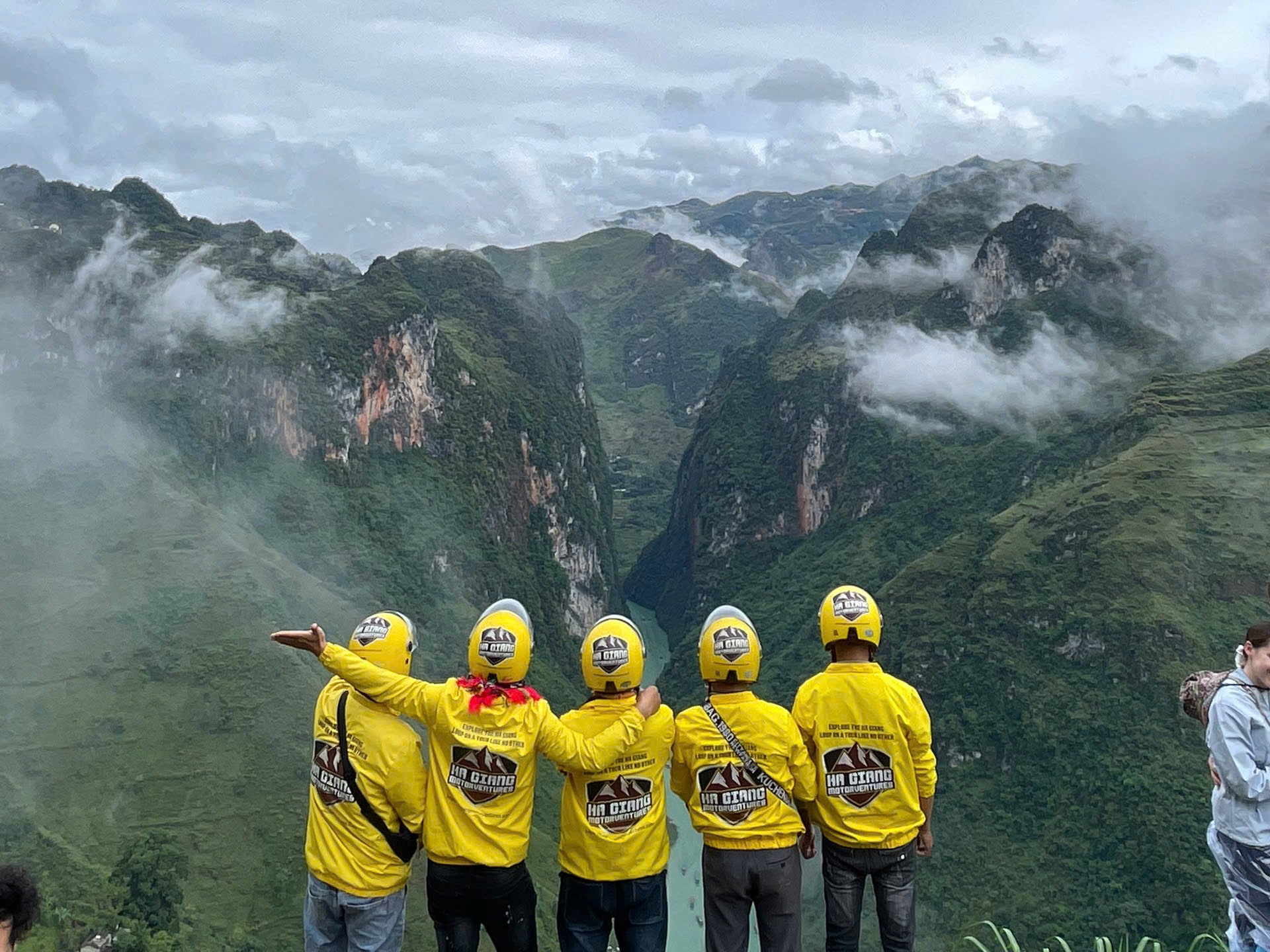 Five Ha Giang Motoventures guides in signature yellow jackets and helmets standing at a misty cliff edge overlooking the Nho Que River, representing our professional tour pickup and drop-off team.