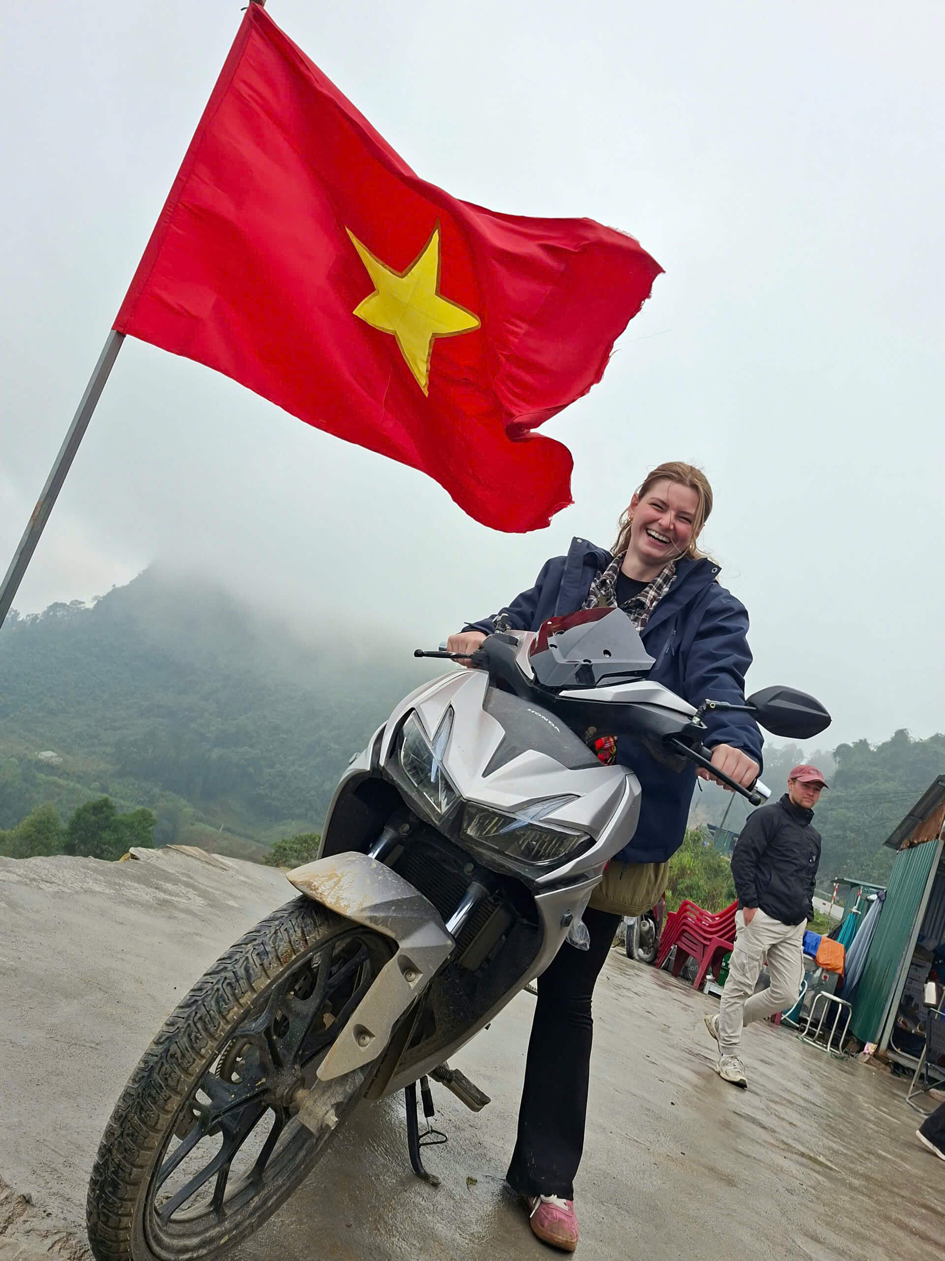 Happy female traveler sitting on a motorbike under the Vietnam flag on a misty day during a Ha Giang Loop adventure