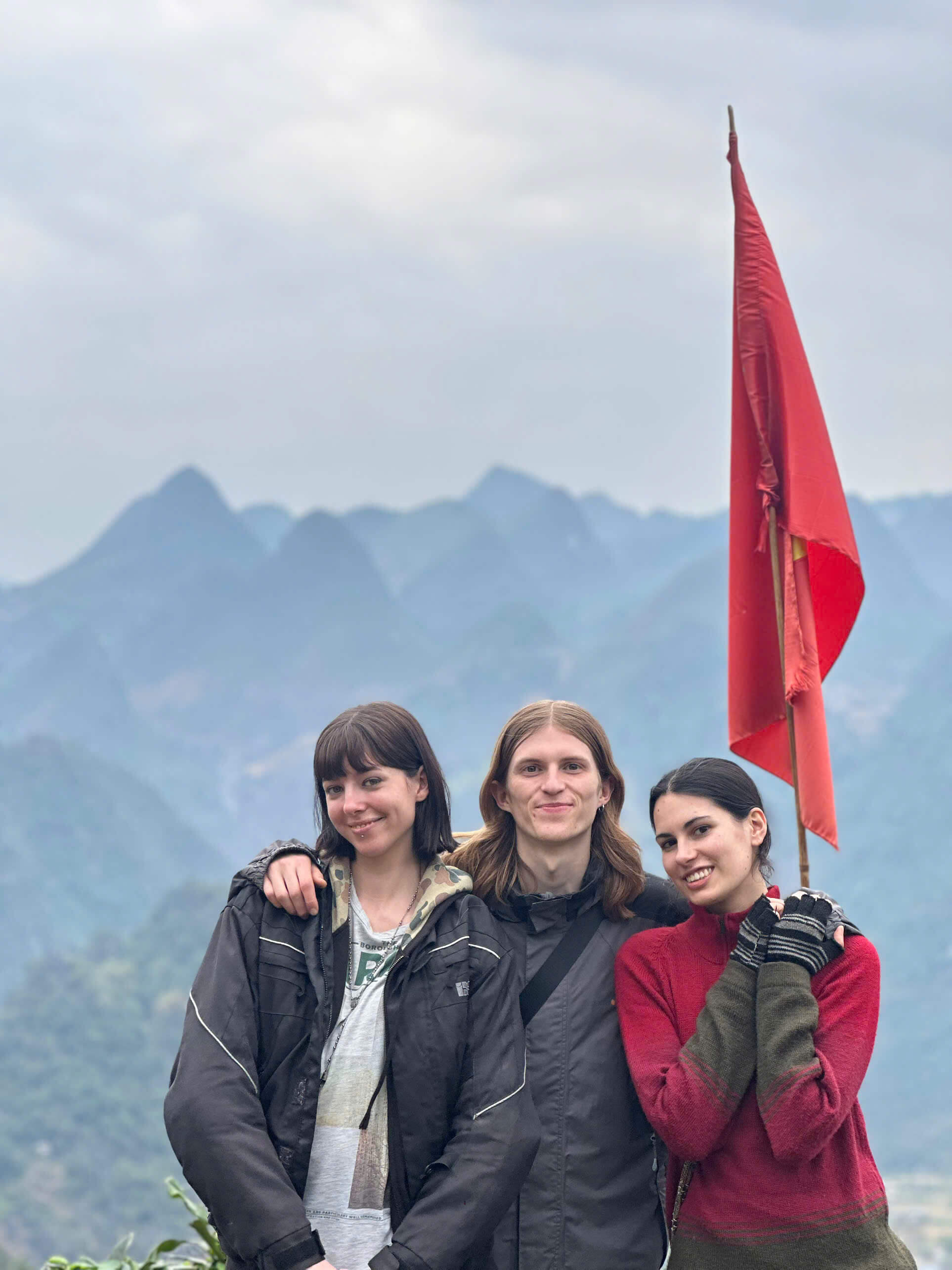 Three travelers posing with the Vietnam flag against a beautiful backdrop of misty karst mountains in Ha Giang