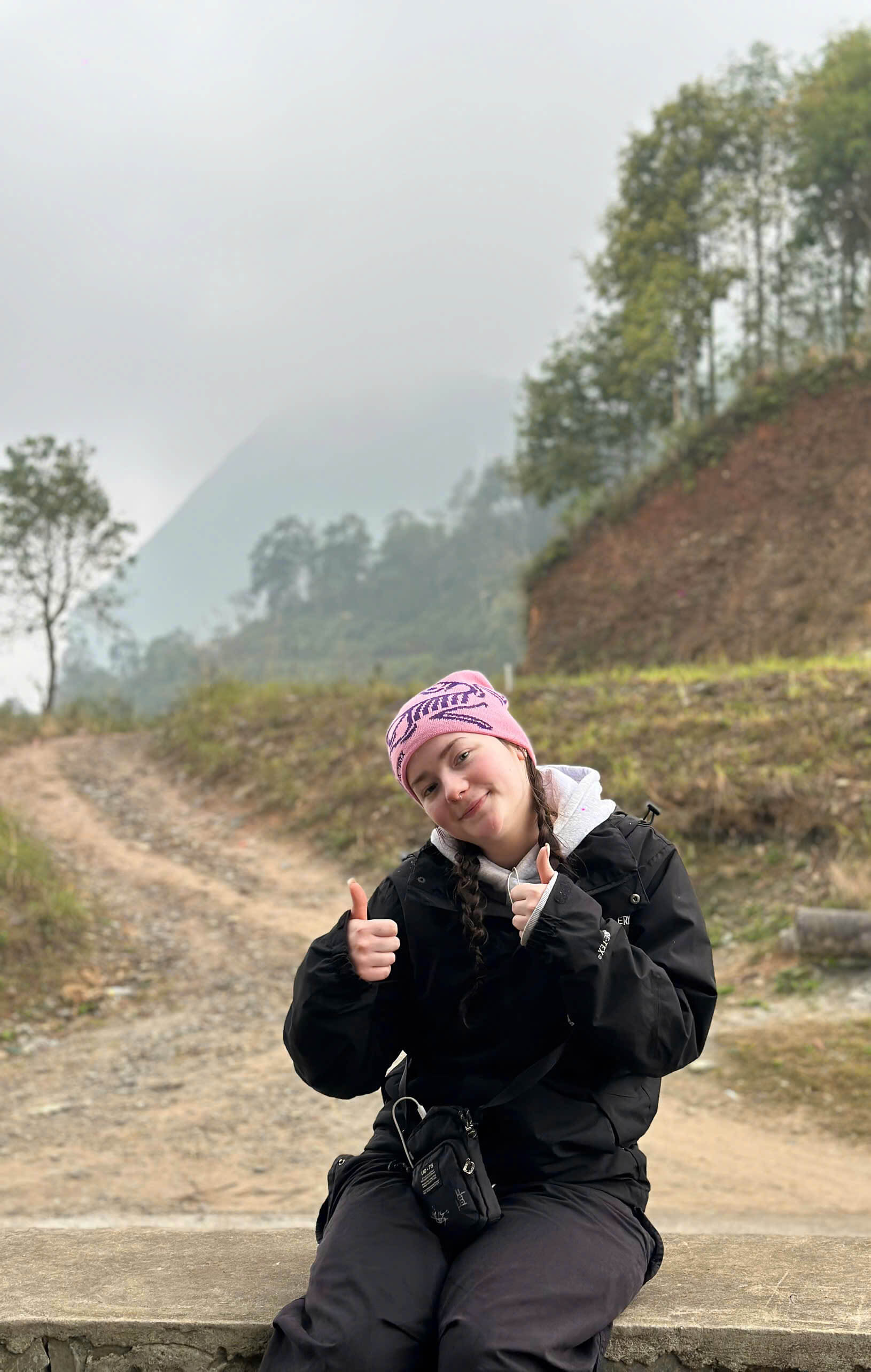 A female backpacker giving a thumbs up while resting on a misty off-the-beaten-path mountain road in Northern Vietnam.