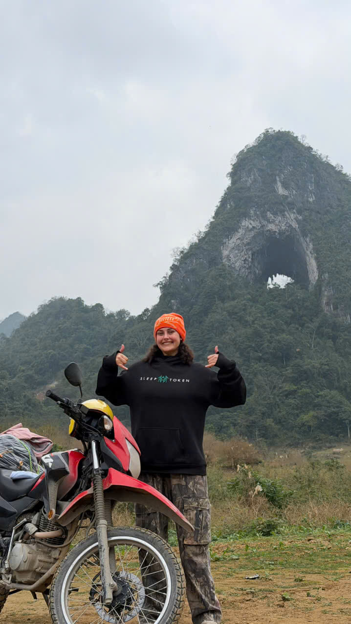 A smiling female traveler posing with her motorbike in front of Angel Eye Mountain on the ultimate Cao Bang Loop adventure.