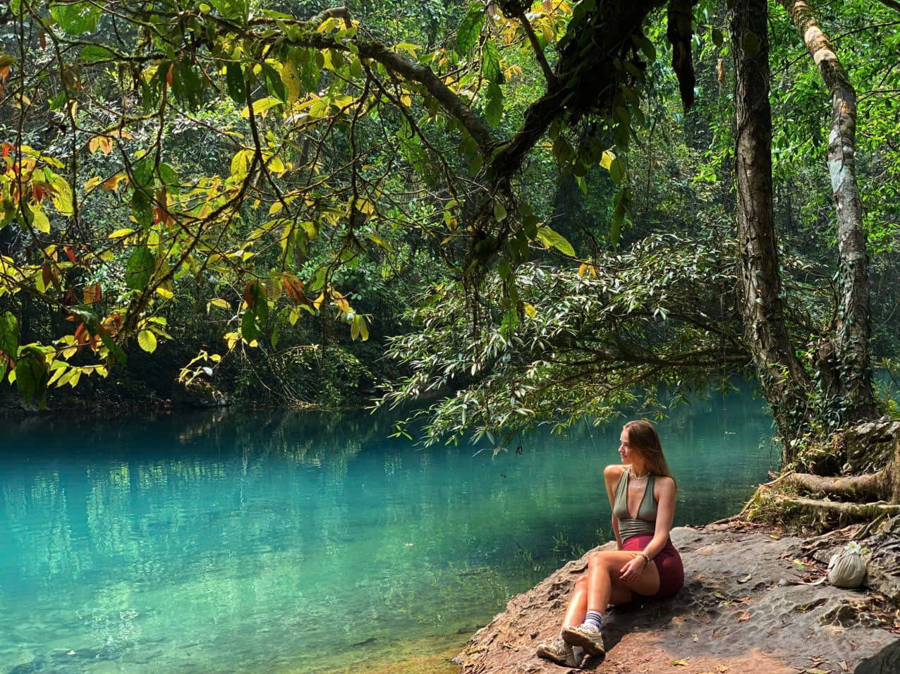 A female traveler sitting peacefully on a rocky bank beside the incredibly clear, turquoise waters of the Lenin River (Suối L&ecirc;-nin) at the Pac Bo historical site. She is surrounded by a lush, green jungle canopy with dappled sunlight reflecting off the emerald water, highlighting the serene and untouched beauty of the Cao Bang Loop.