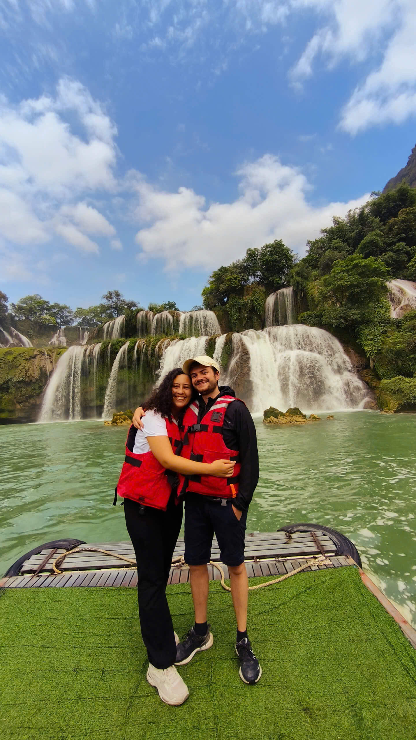 A happy couple in life jackets hugging in front of the majestic Ban Gioc Waterfall during their Cao Bang Loop tour.