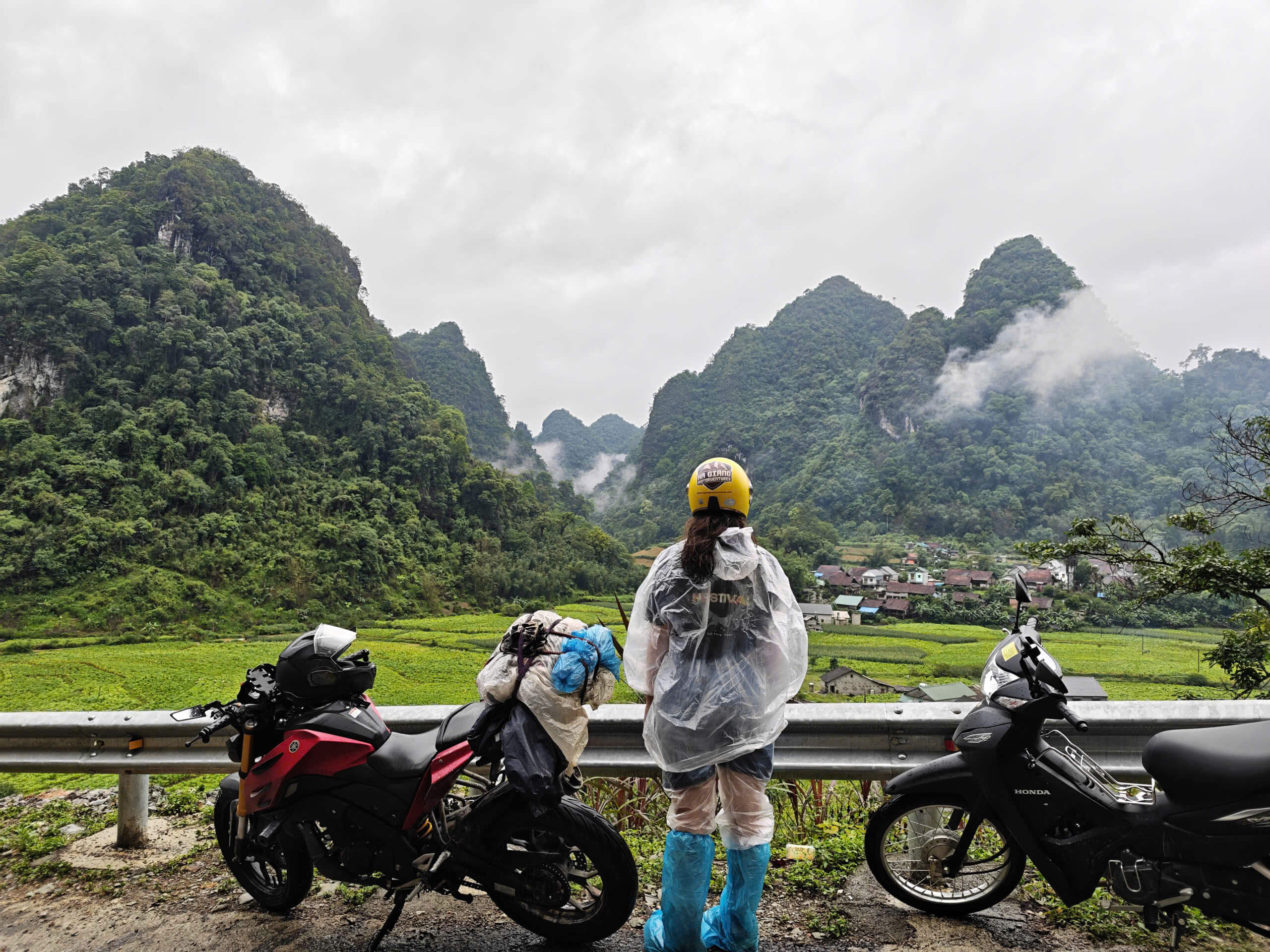 A traveler wearing a rain poncho stands beside their loaded motorbikes, gazing out over a breathtaking, misty green valley surrounded by towering limestone mountains on the Cao Bang Loop.