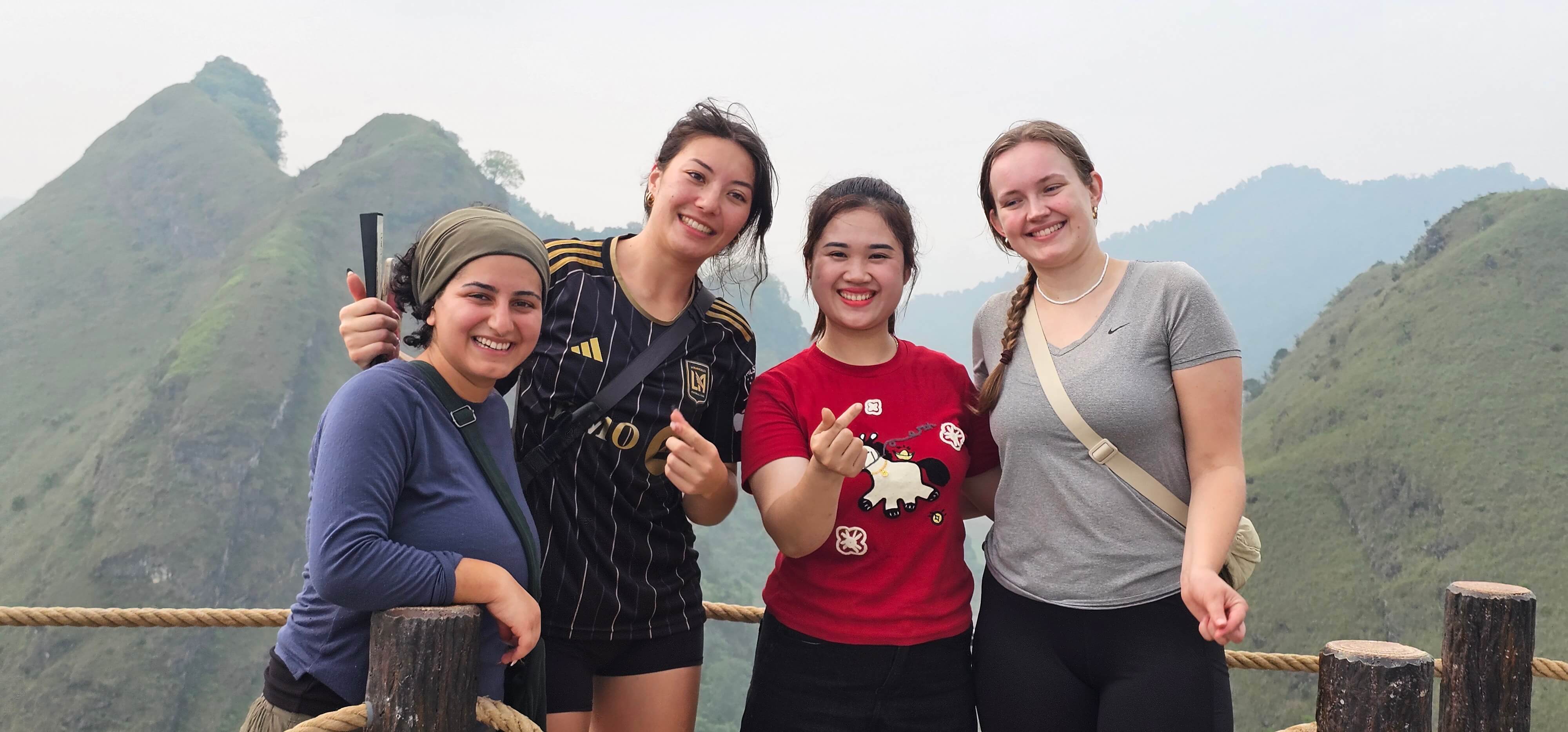 Happy travelers taking a group photo on a scenic mountain road in Cao Bang, with large limestone karst cliffs and dense green foliage in the background during a motorbike tour.