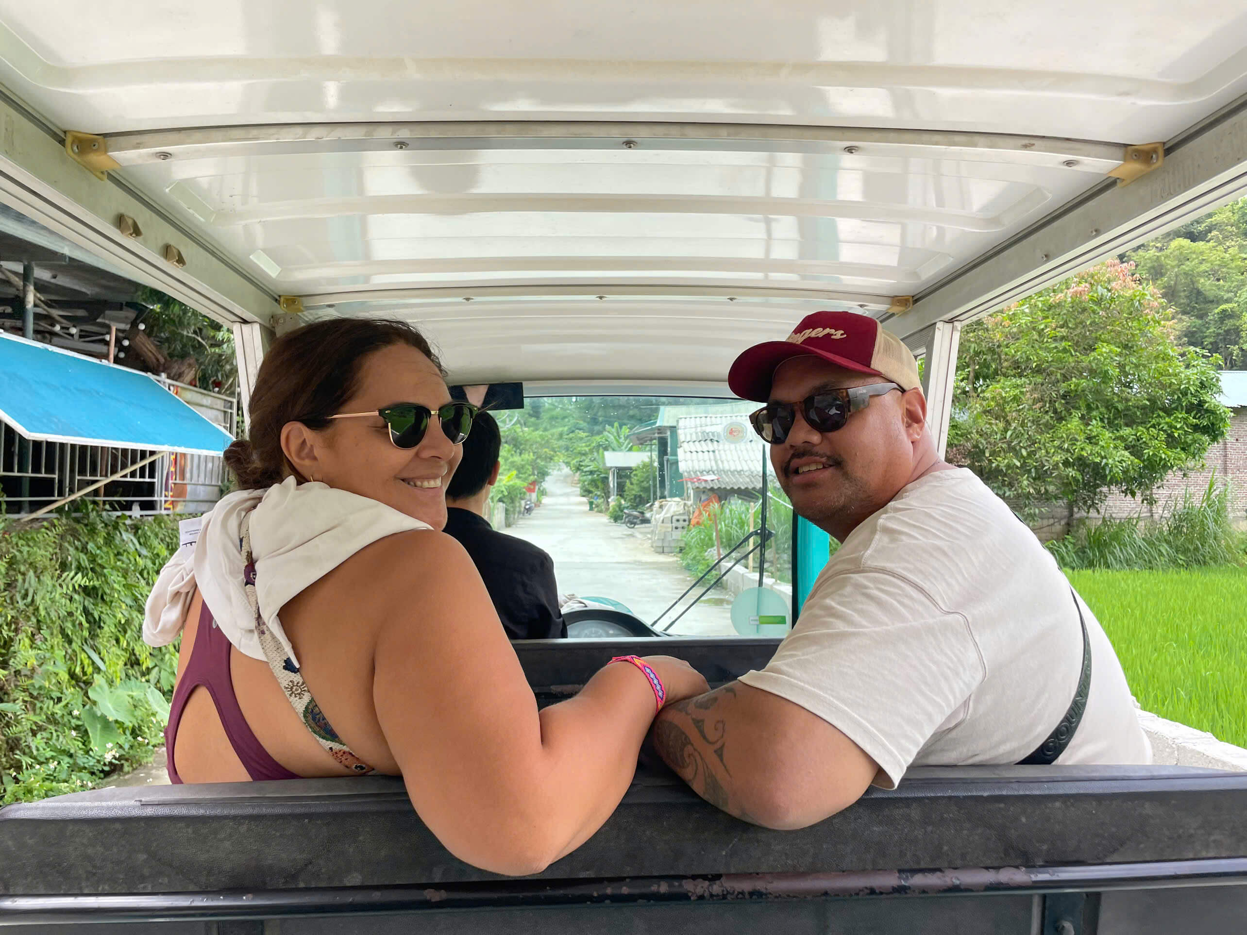 A smiling couple looking back while being chauffeured in a buggy-like transport vehicle through a local village in Cao Bang, offering an alternative to a traditional motorbike tour for couples with Motorventures.
