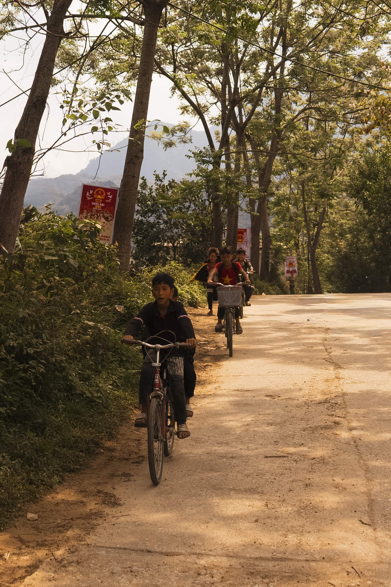 Local ethnic minority children riding bicycles down a sunlit rural road, showcasing the authentic everyday village life encountered on a Motorventures tour.