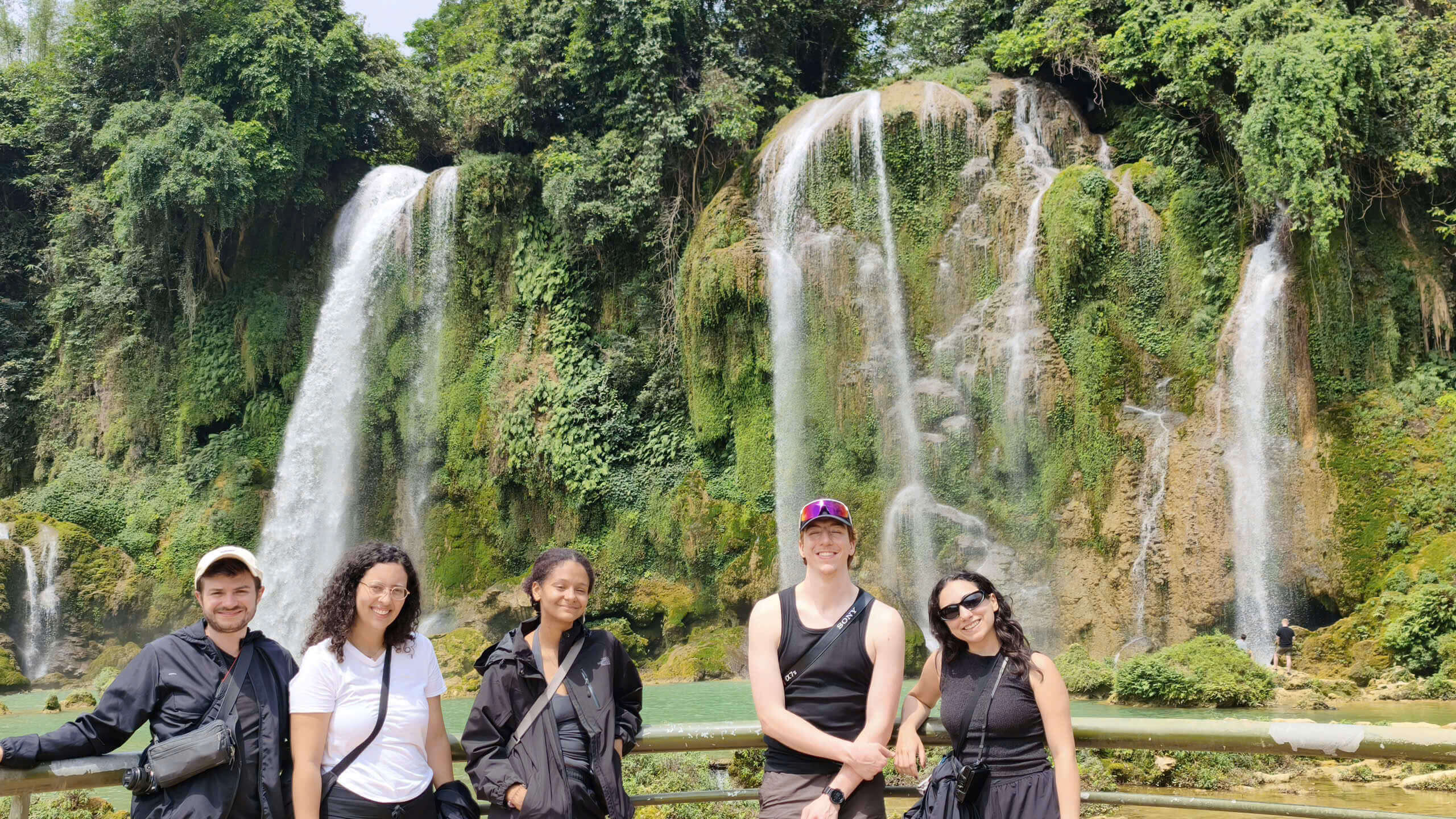 A group of five happy international travelers posing in front of the spectacular, multi-tiered Ban Gioc Waterfall during their Cao Bang motorbike loop tour with Motorventures.
