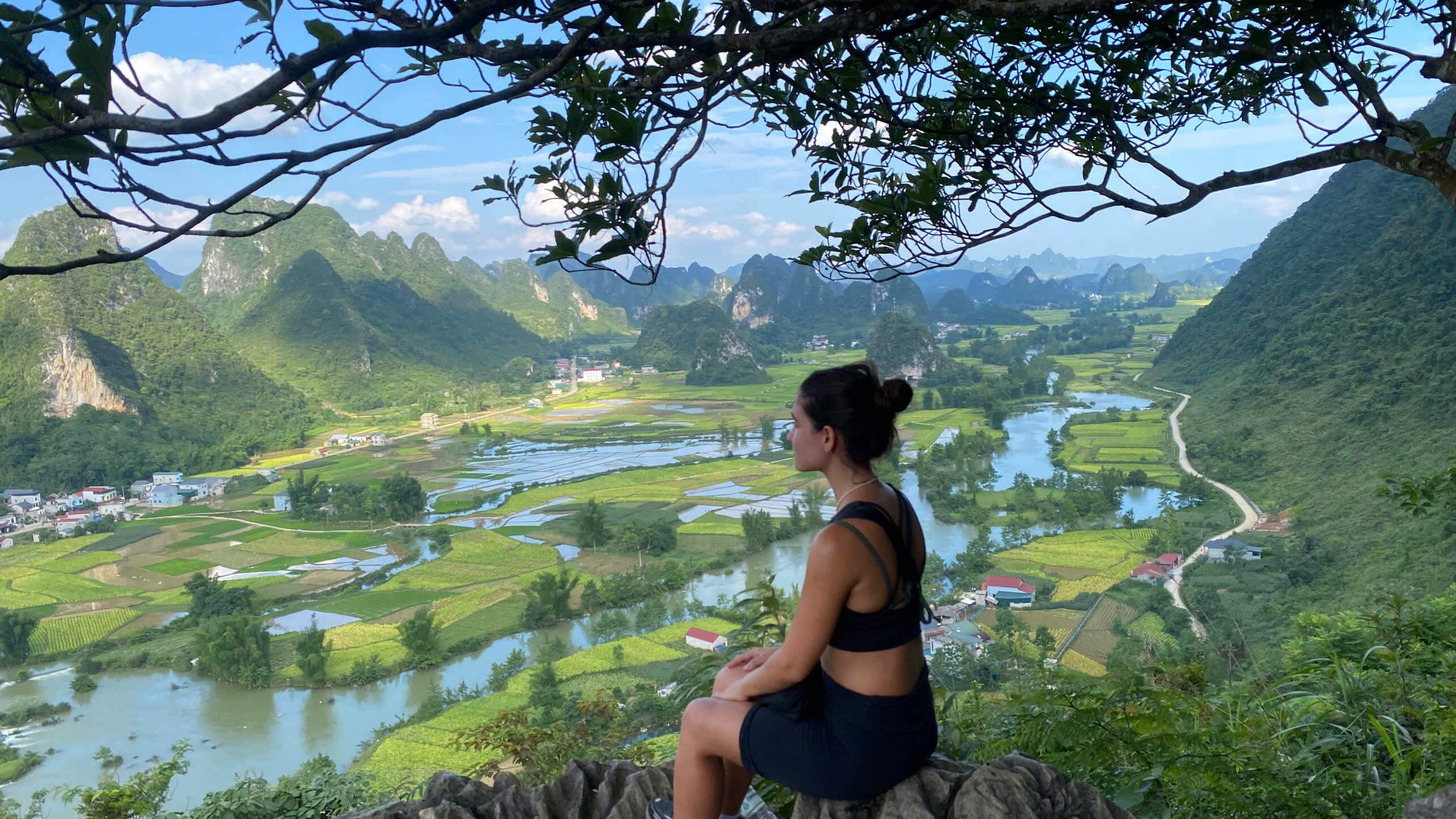 A female traveler sitting on a rocky viewpoint, admiring the stunning landscape of a winding river, lush green rice paddies, and limestone karsts in a Cao Bang valley during a motorbike tour.