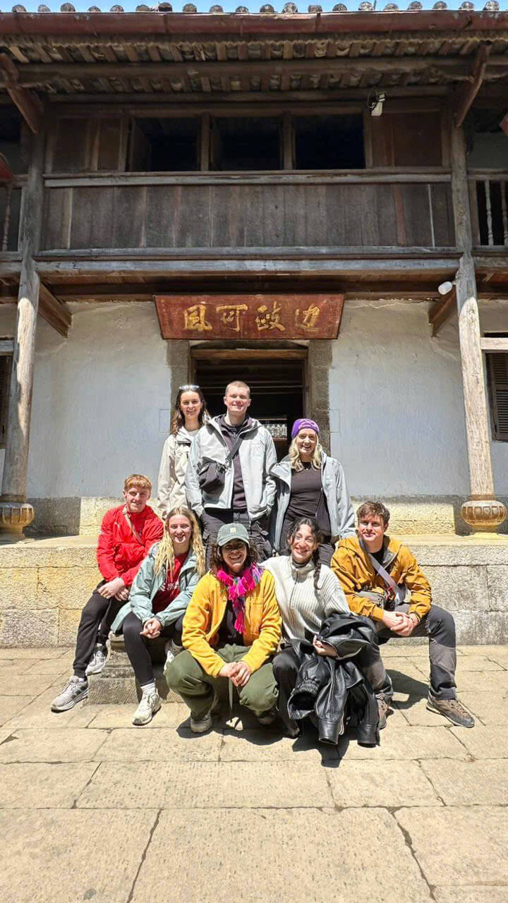 A smiling group of eight Motorventures travelers posing together on the stone steps of the historic Hmong King's Palace (Dinh Vuong) during their Ha Giang loop cultural itinerary.