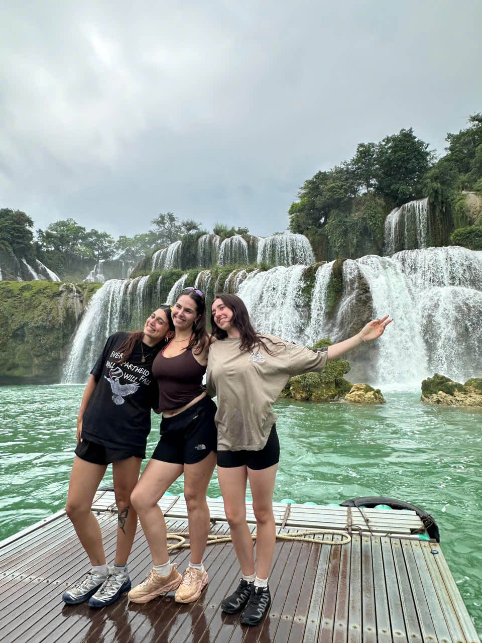 Three happy female tourists standing on a wooden raft on the emerald river, getting a breathtaking up-close view of the thundering Ban Gioc Waterfall in Cao Bang.