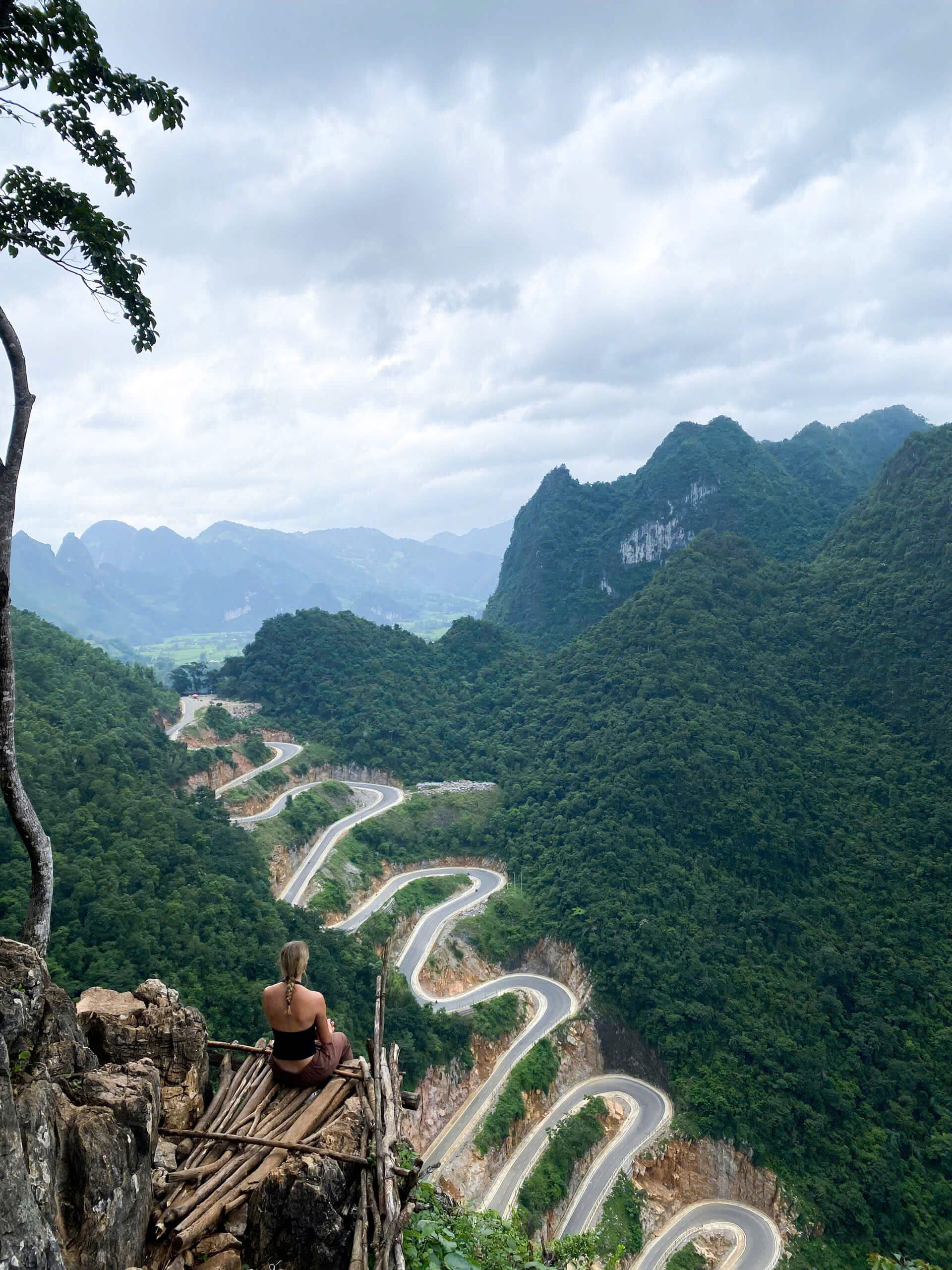 A female traveler sitting on a rustic wooden platform on a cliff rim, looking down at the spectacular 14-tier Khau Coc Cha winding mountain pass during a Cao Bang motorbike loop.