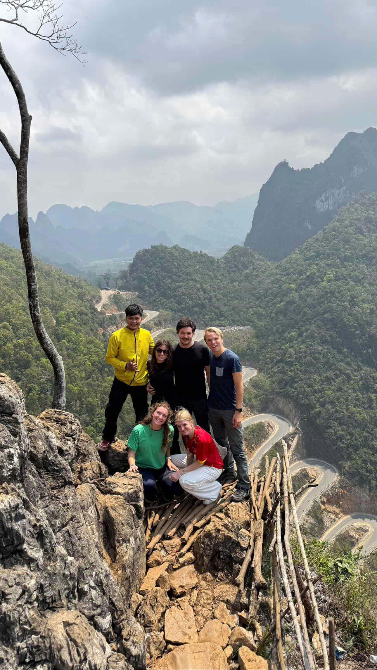 A group of Motorventures travelers and their local guide posing safely on a rocky cliff overlooking the spectacular winding 14-tier Khau Coc Cha pass on the Cao Bang loop.