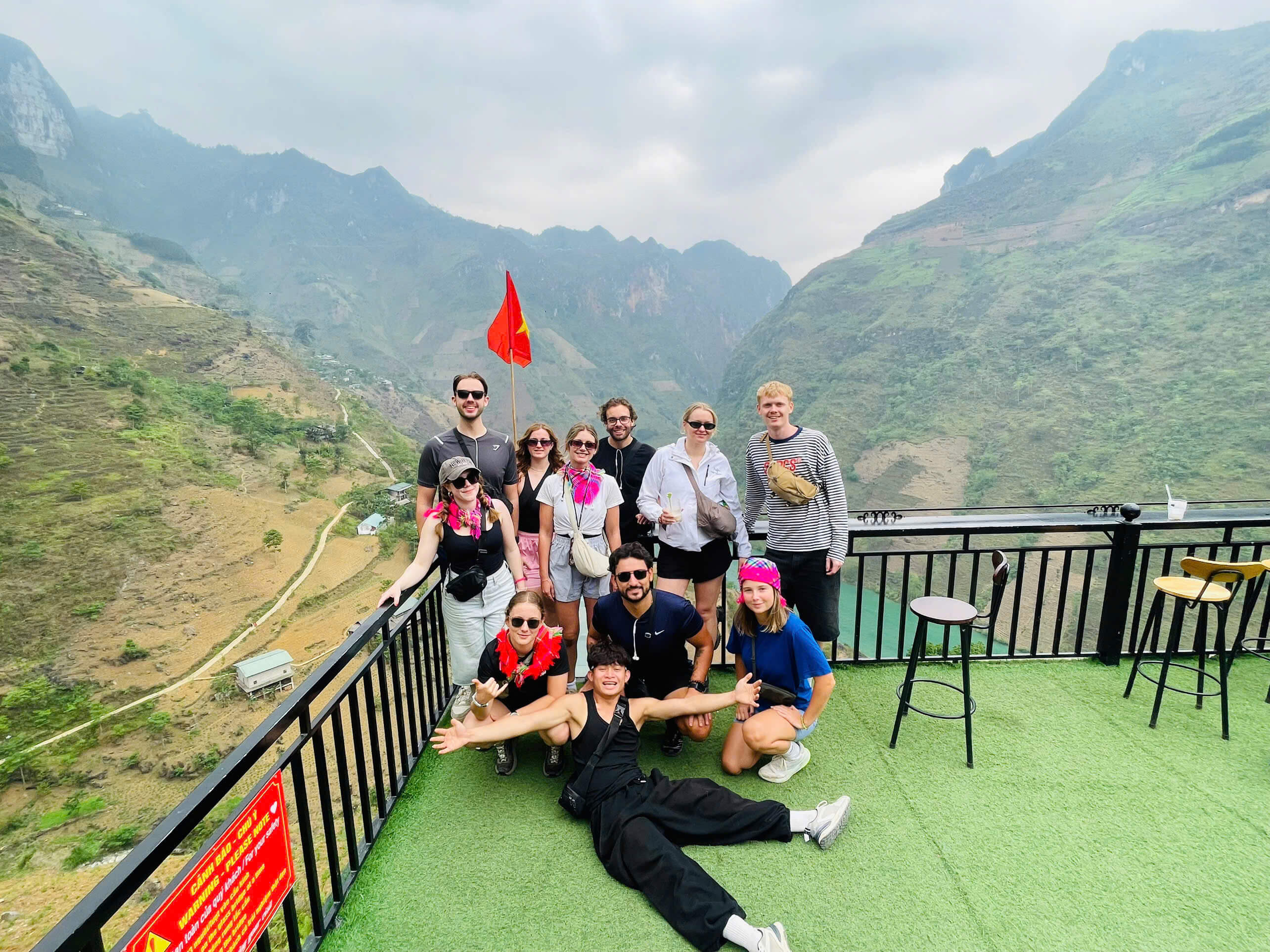 A cheerful group of international tourists posing playfully on a green viewing deck overlooking the magnificent Nho Que River canyon during a Ha Giang loop tour.