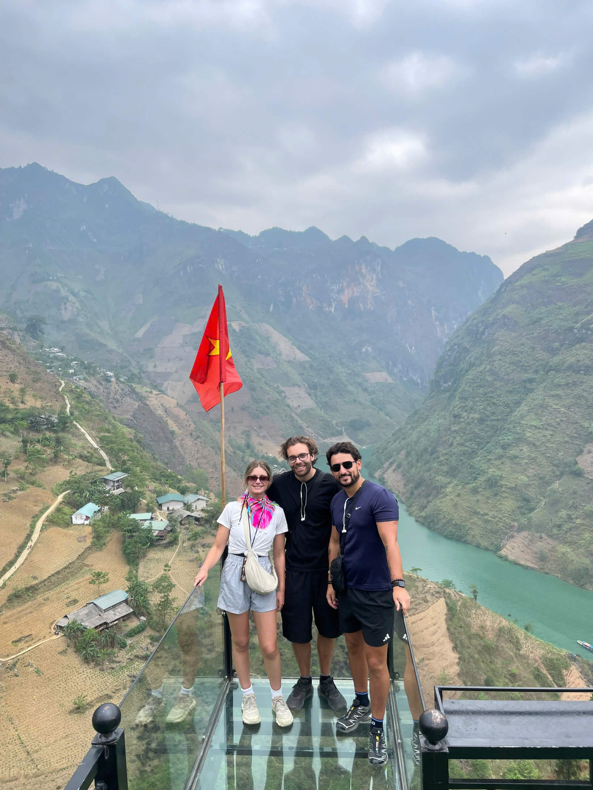 Three smiling travelers standing on a transparent glass balcony beside a Vietnamese flag, enjoying the breathtaking high-altitude view of Tu San Canyon and the emerald river below.