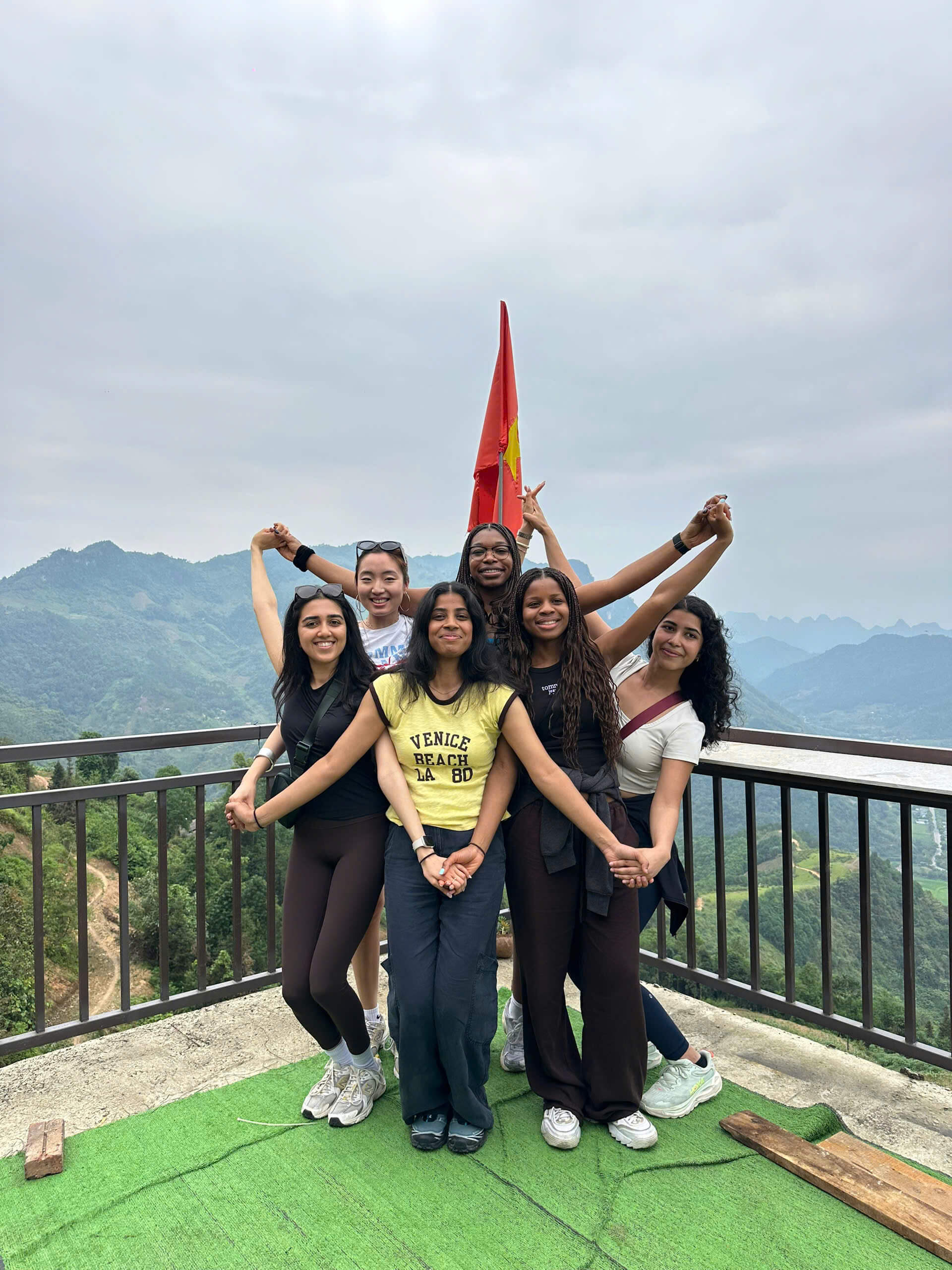 A group of happy female travelers holding hands and posing cheerfully in front of a Vietnamese flag at a high-altitude mountain viewpoint during their Ha Giang loop tour.