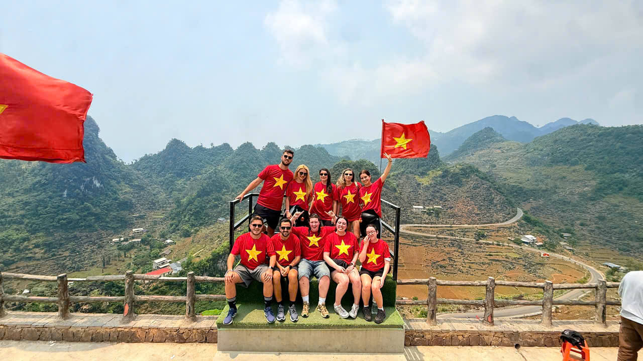 A large, smiling group of Motorventures travelers wearing matching red Vietnamese flag t-shirts, sitting on a viewing deck overlooking the iconic winding roads of the Ha Giang loop.