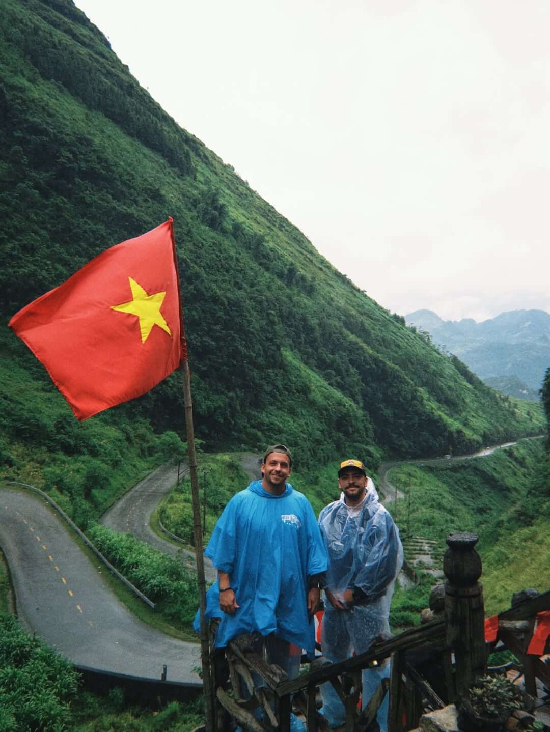 Two male travelers wearing rain ponchos posing next to a Vietnamese flag at the famous Tham Ma Pass viewpoint, showcasing the iconic winding mountain roads in the background.