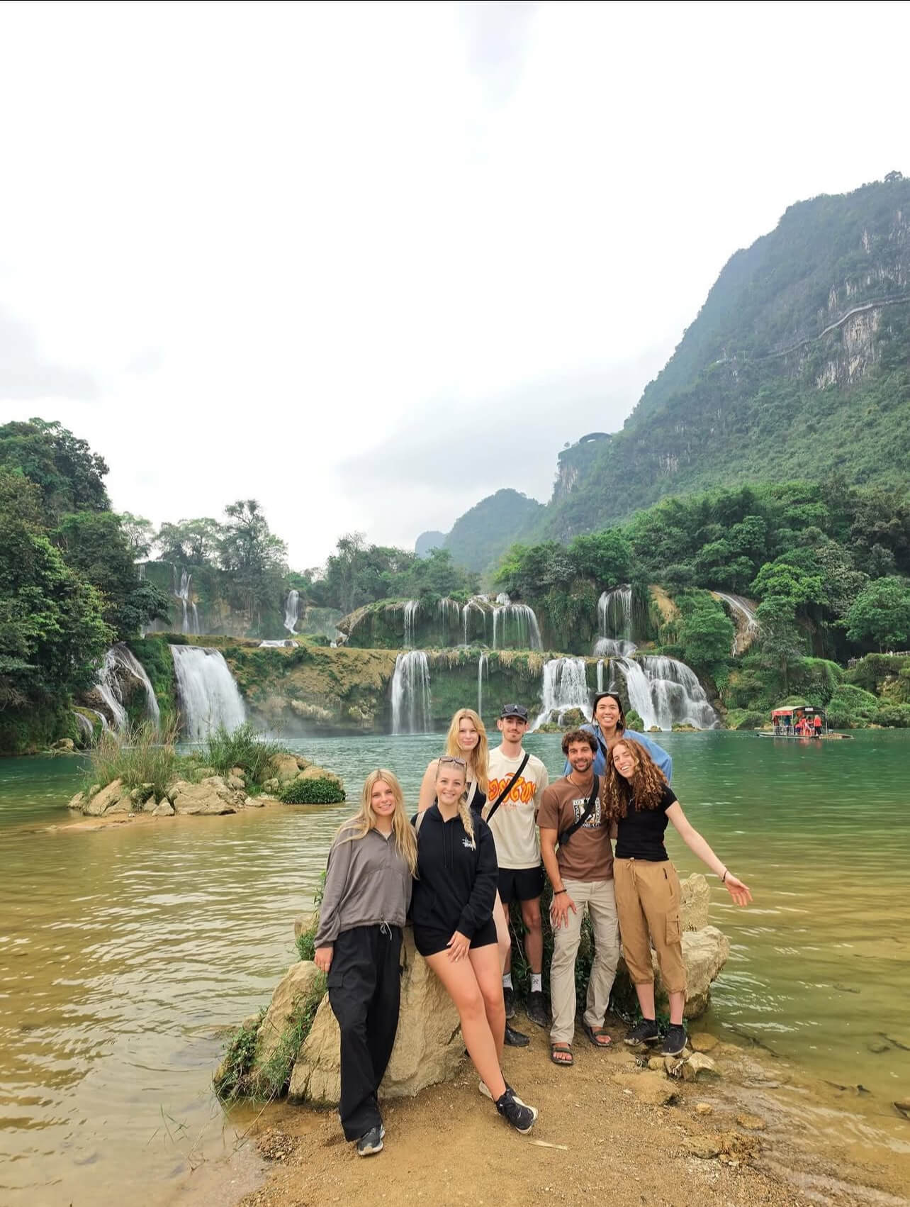 A group of seven young travelers smiling and posing together on a small rock island in the emerald river, with the massive, multi-tiered Ban Gioc Waterfall roaring in the background.