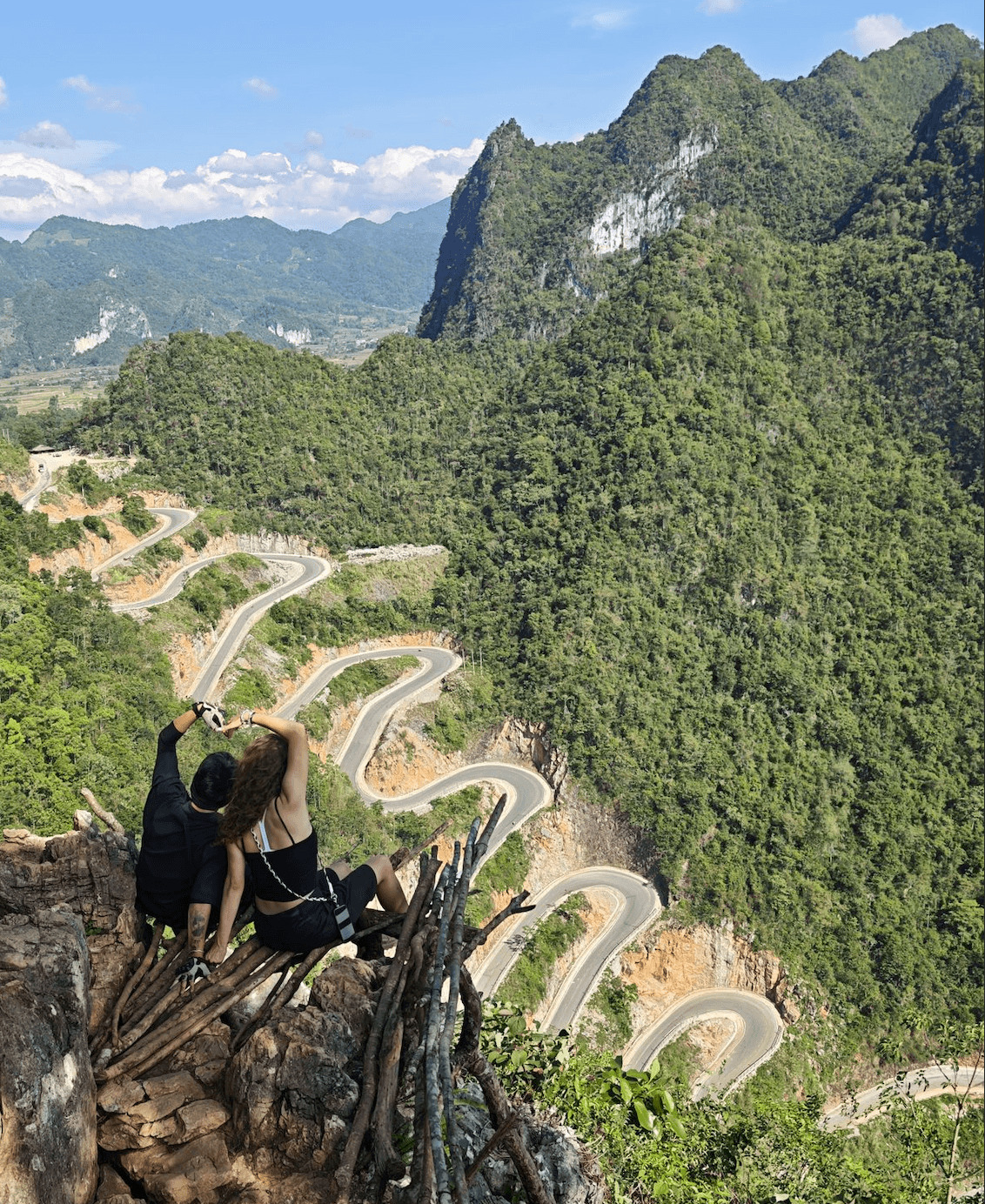 A couple sitting together on a rustic cliffside viewpoint, making a heart shape with their arms while overlooking the winding 14-tier Khau Coc Cha mountain pass on the Cao Bang Loop.
