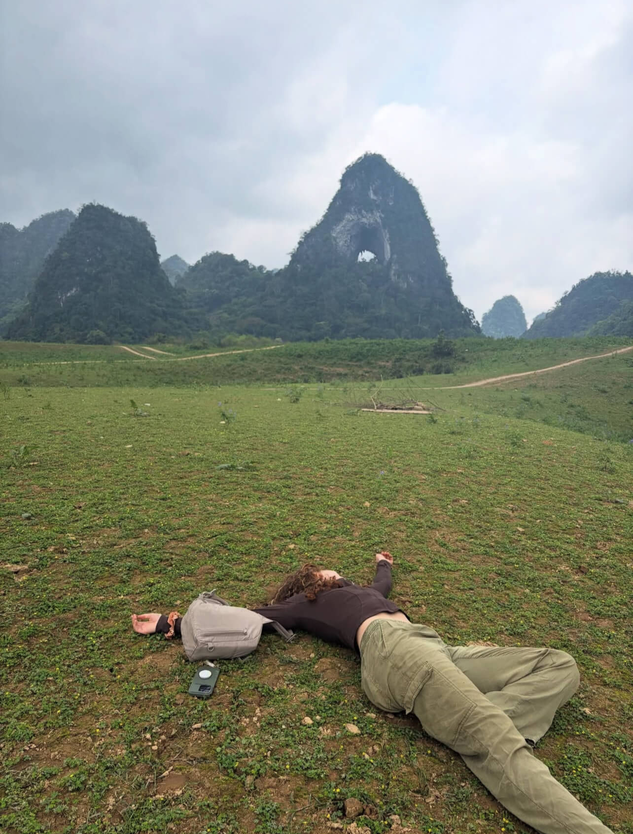 A female traveler lies spread-eagle and relaxed on a vast, green grassy plain, with the unique "Angel Eye Mountain" (Nui Thung)—a karst peak with a large hole through it, in the background.
