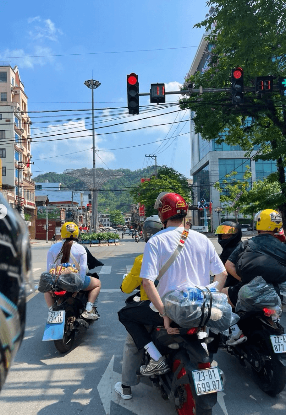 Backpackers and locals riding motorbikes with luggage strapped to the back, waiting at a red traffic light in the modernizing streets of Ha Giang City.