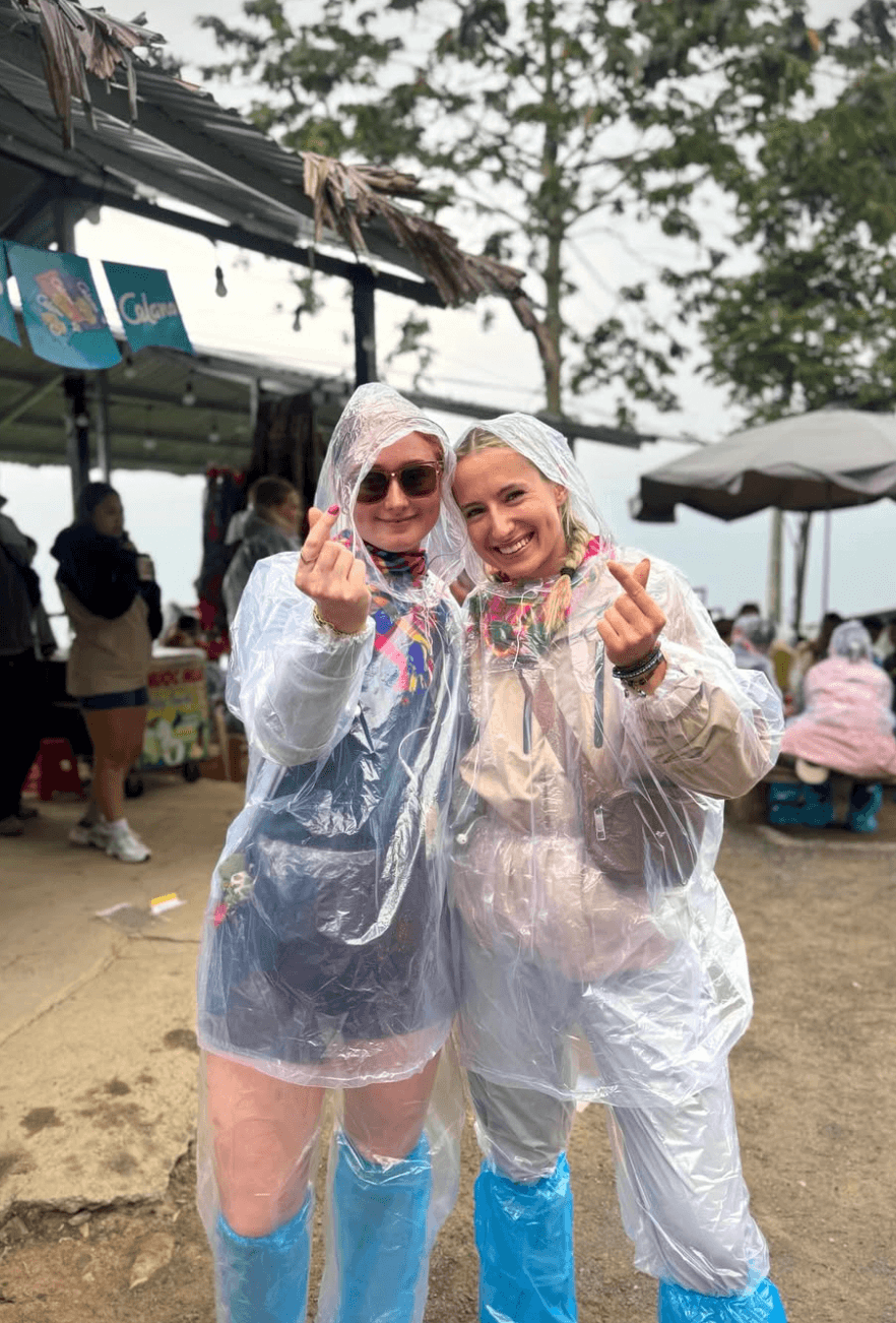 Two happy female travelers smiling and making finger hearts while wearing clear plastic rain ponchos and waterproof blue shoe covers at a mountain rest stop in Ha Giang.