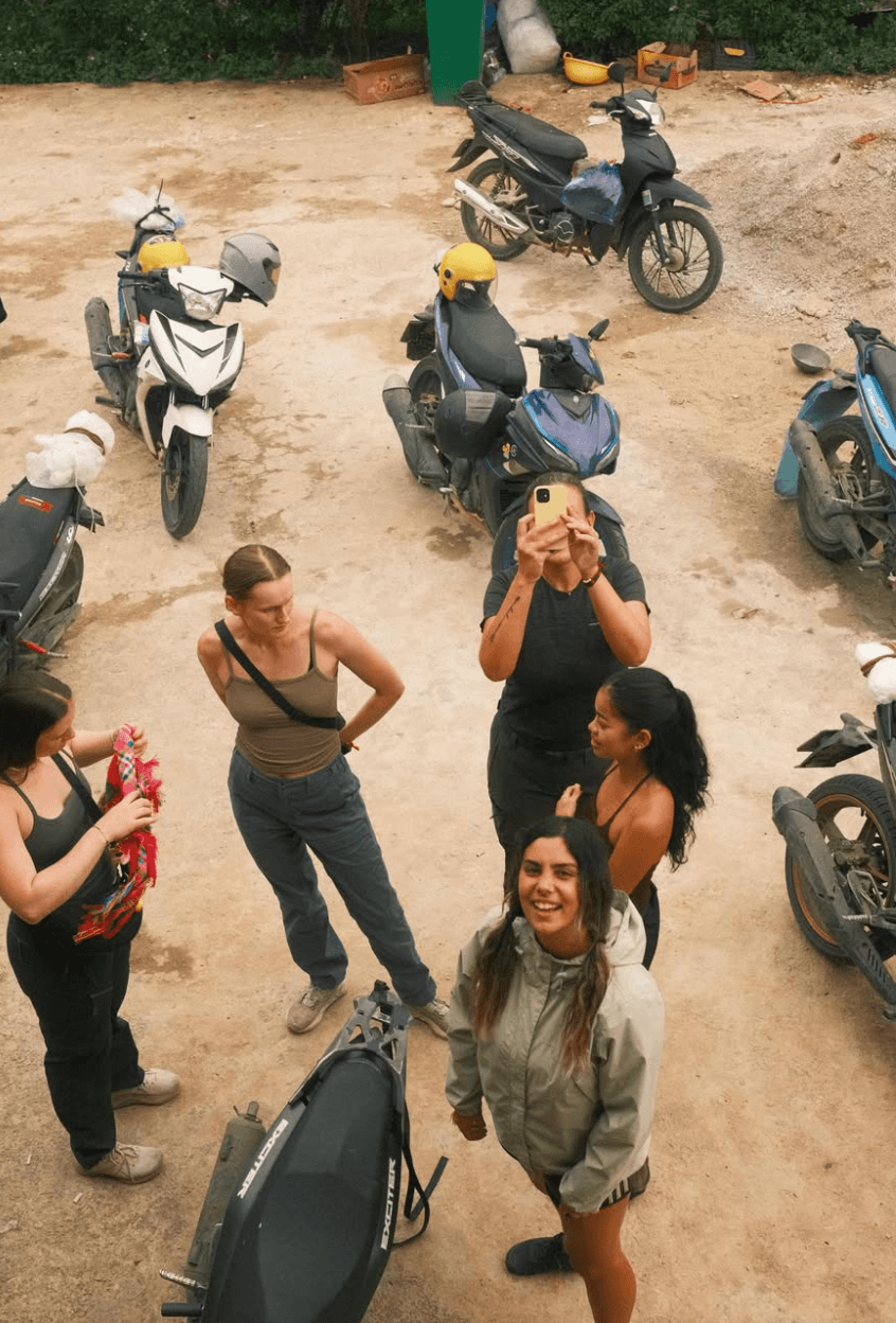 : A group of female tourists taking photos on their phones and relaxing next to a line of parked motorbikes at a dirt rest stop in Ha Giang.
