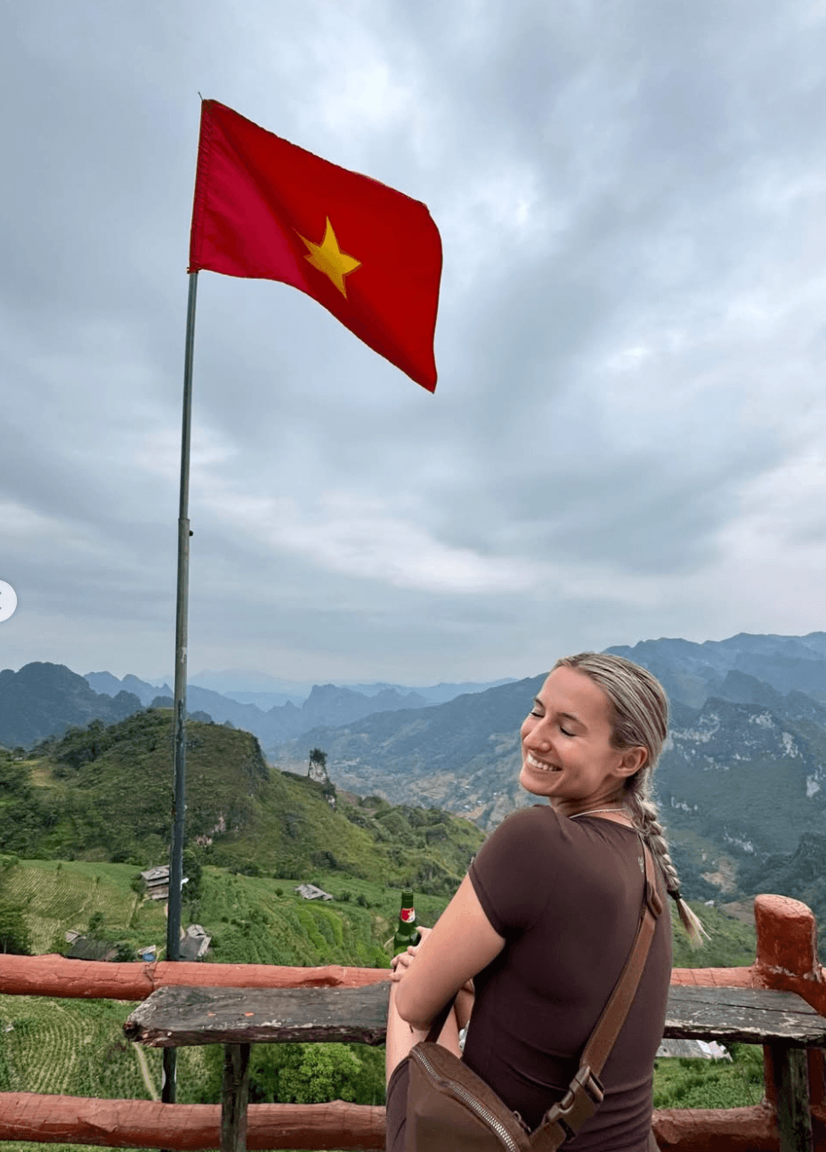 A blonde female traveler smiling peacefully with her eyes closed while sitting at a high-altitude scenic viewpoint, with a large Vietnamese flag waving behind her against a backdrop of sweeping mountains.