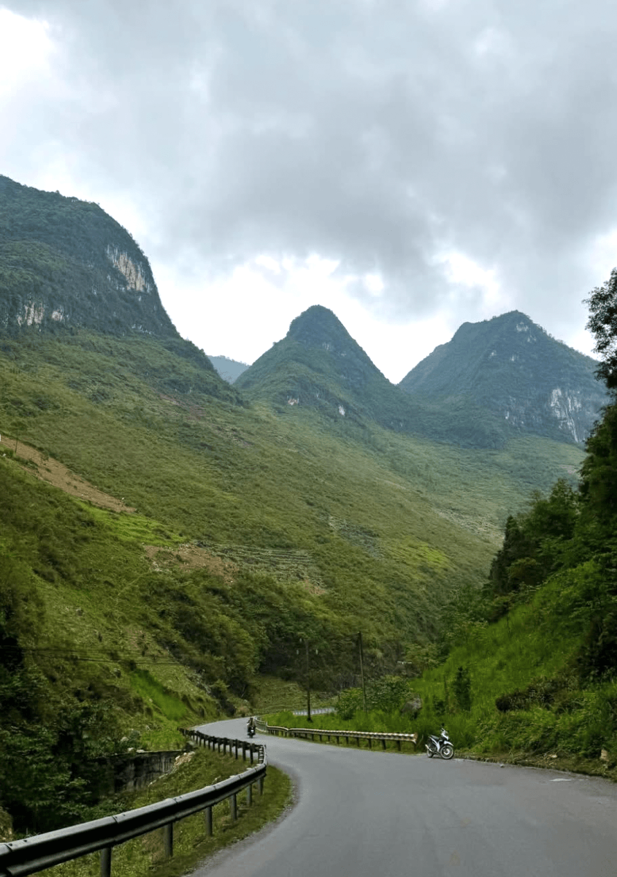 A view of a winding asphalt road hugging the side of a lush green mountain on the Ha Giang Loop, with towering limestone karst peaks in the background under a cloudy sky.