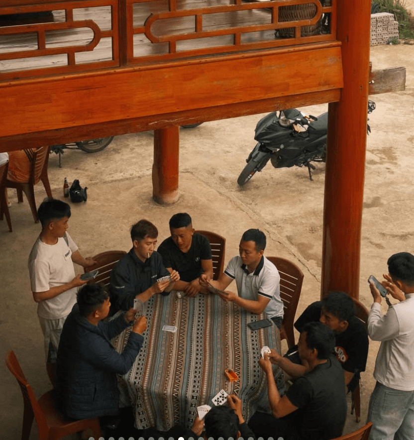 A group of local Vietnamese men, working as Easy Rider guides, relaxing and playing a card game around a table beneath a traditional wooden stilt house in Ha Giang.