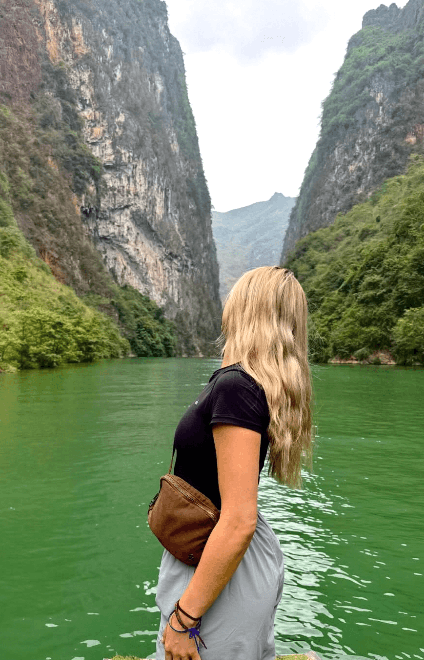 A female traveler wearing a breathable black short-sleeve t-shirt, lightweight grey pants, and a small brown crossbody daypack, looking up at the towering Tu San Canyon on the Ha Giang Loop.