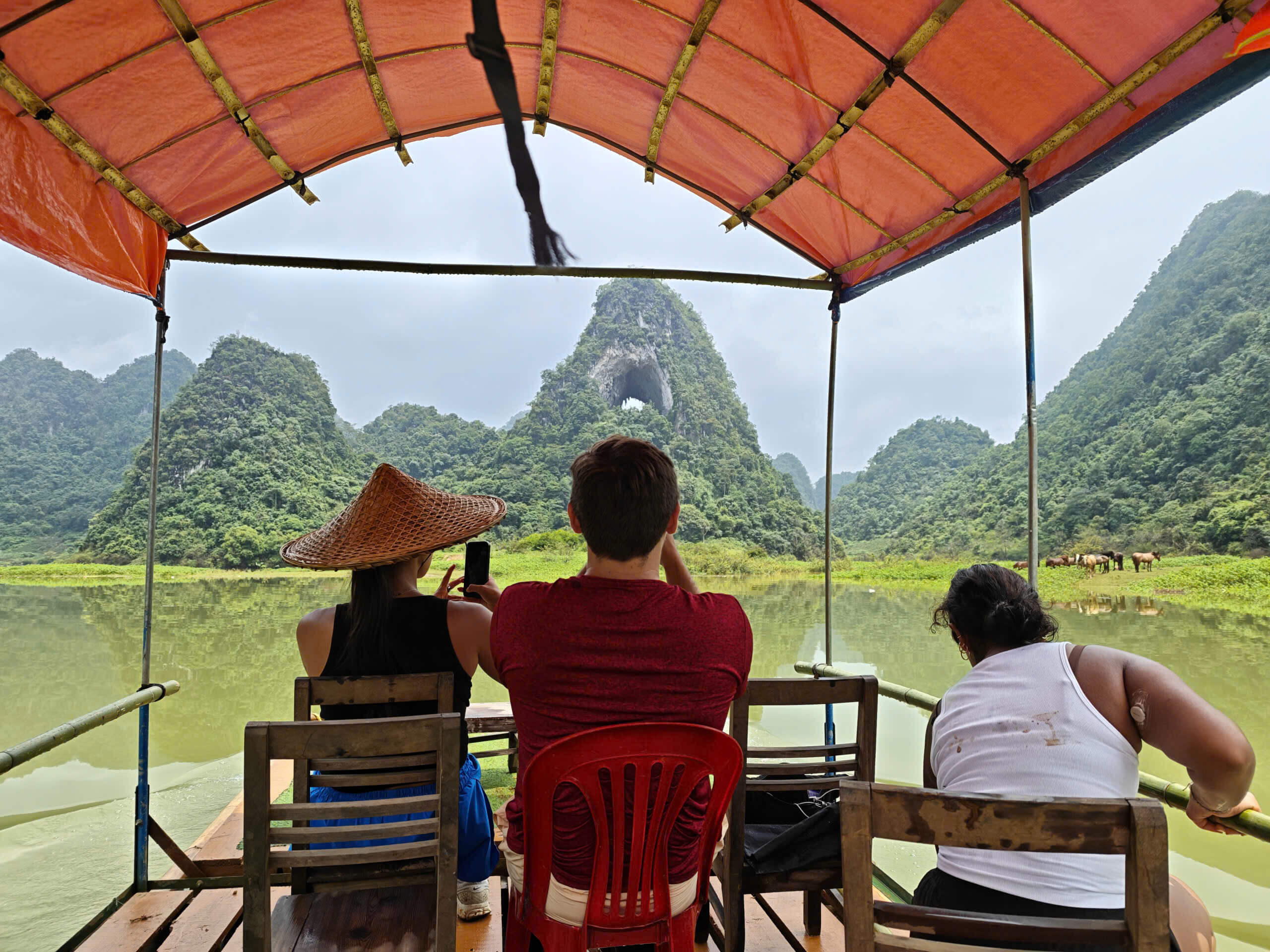 A male tourist and a local Motorventures guide sitting together on the edge of a green raft, looking at a smartphone, with Angel Eye Mountain looming in the background.