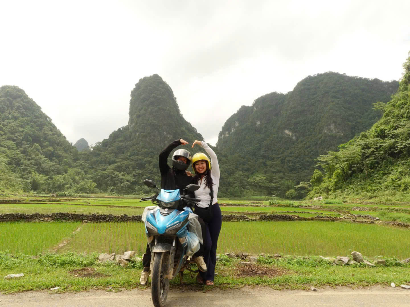 A couple sitting on a blue motorbike making a heart shape with their arms over their helmets, surrounded by vibrant green rice paddies and limestone karst mountains on the Cao Bang loop.