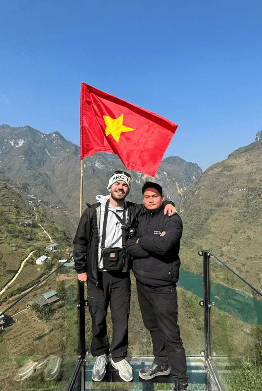 A tourist riding pillion on a motorbike playfully makes a heart shape with their hands over their Easy Rider guide's helmet while cruising down a scenic, rugged mountain road in Ha Giang.