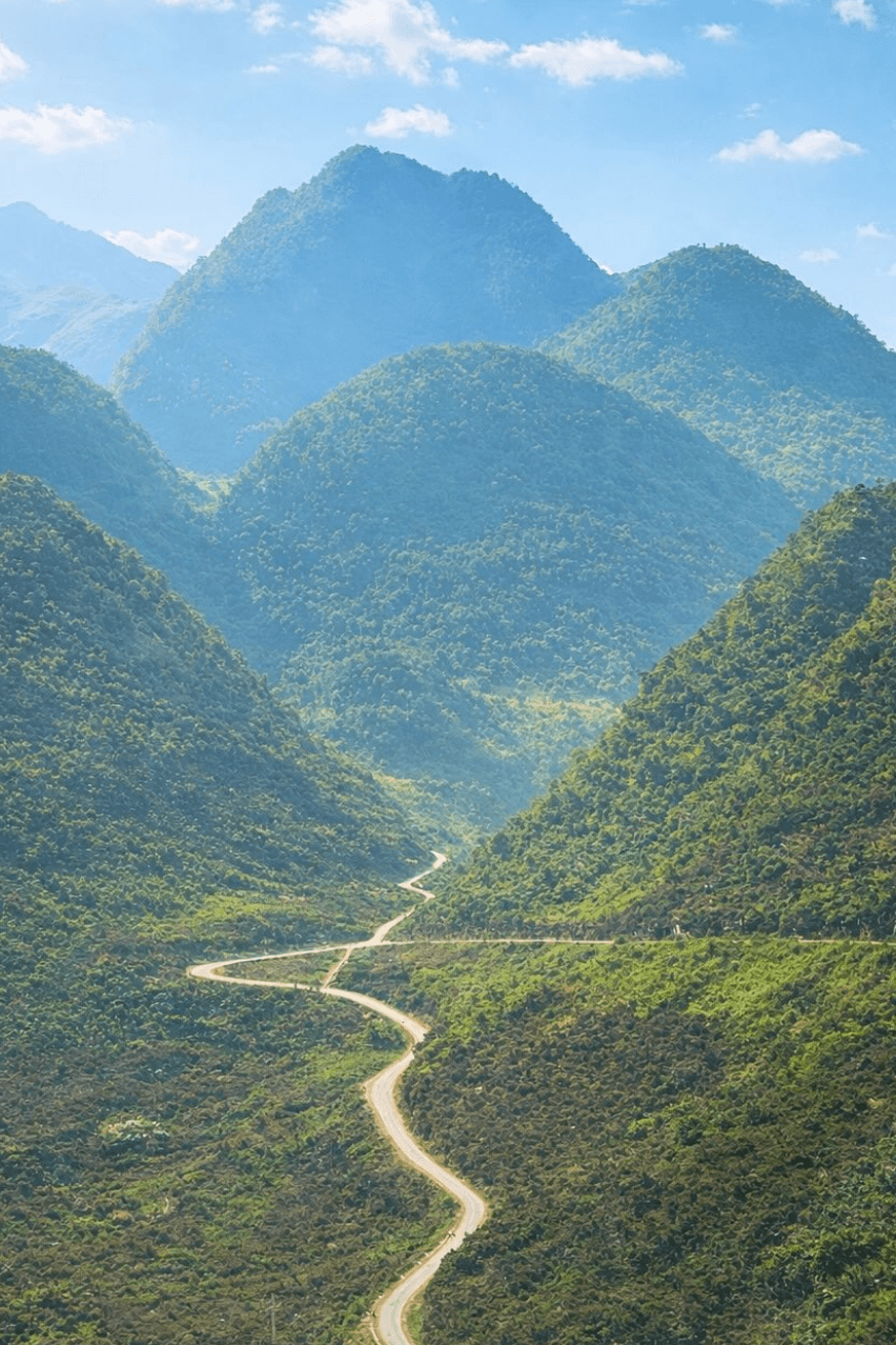 A breathtaking landscape view of a winding, narrow paved mountain road cutting through a deep, lush green valley surrounded by towering, layered karst peaks under a clear blue sky in Northern Vietnam.