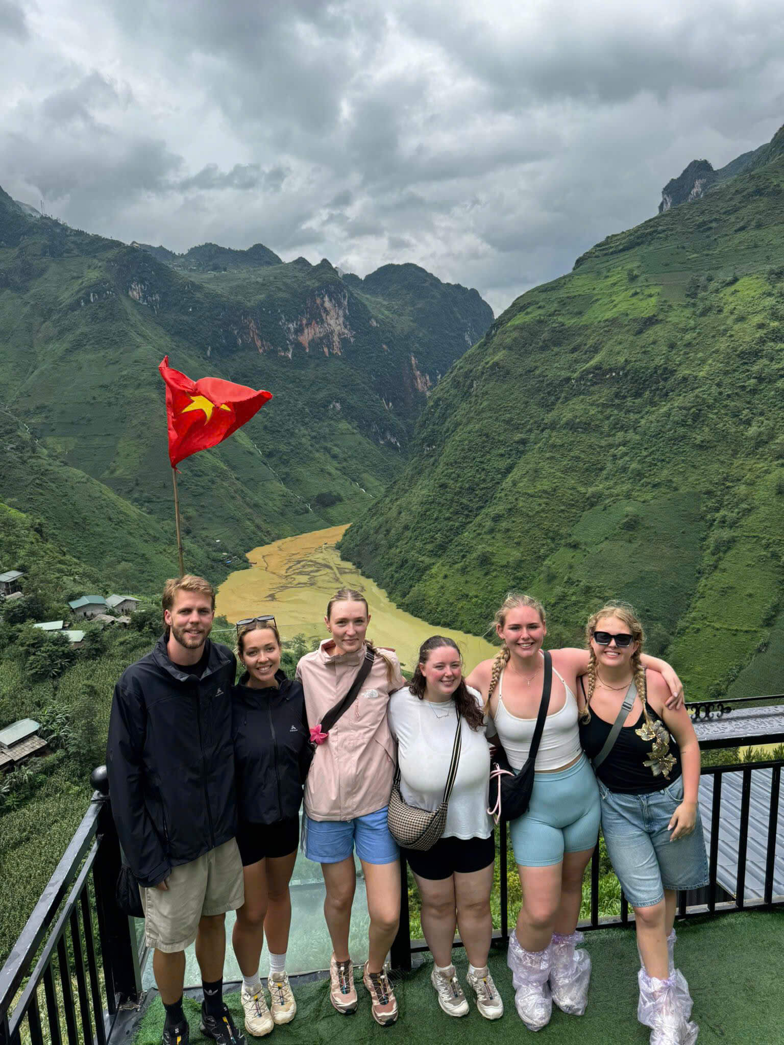 A smiling group of tourists wearing orange life jackets and rain ponchos taking a boat ride through the towering, misty Tu San Alley canyon on the Nho Que River in Ha Giang.