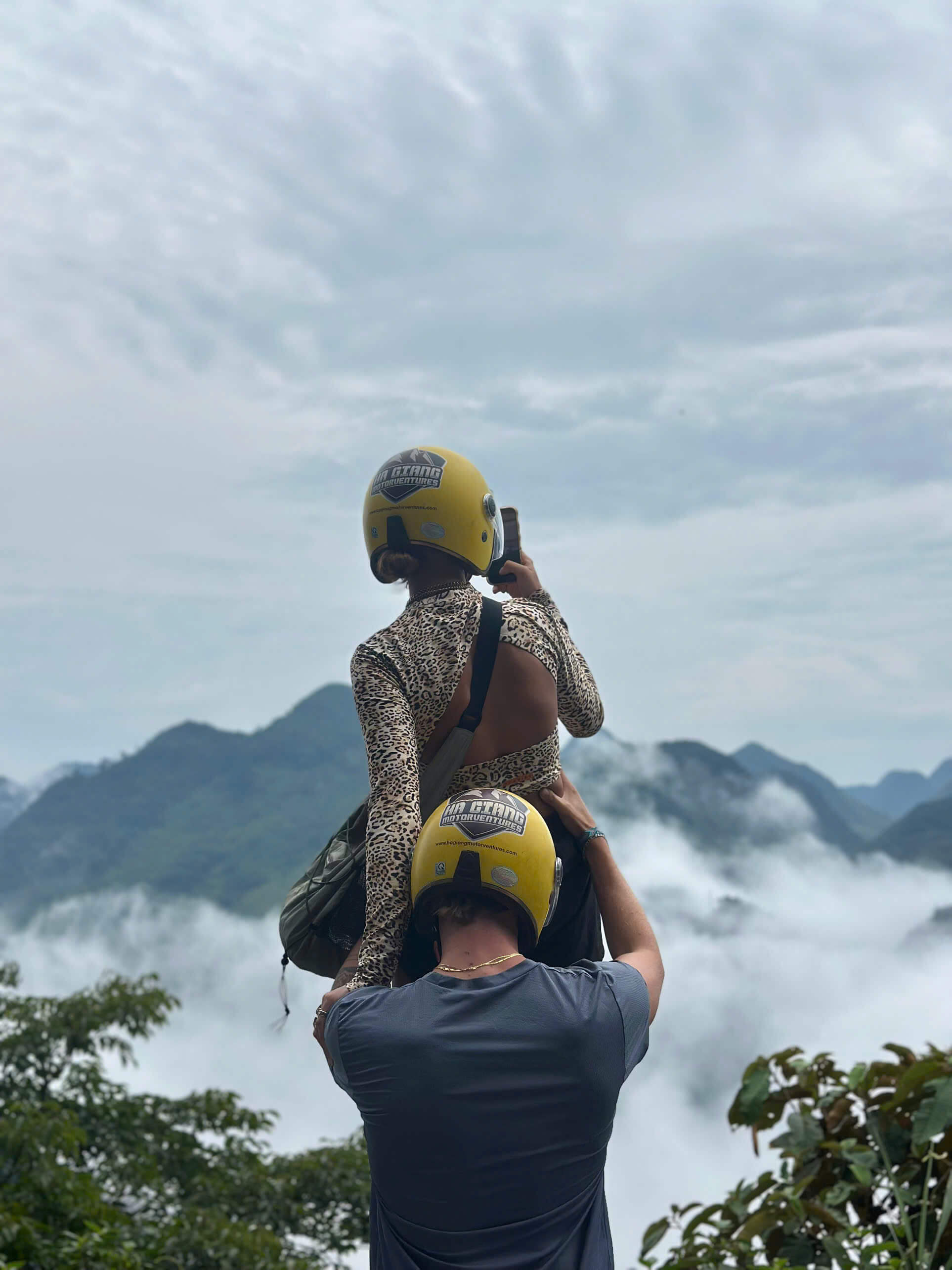A man wearing a yellow Ha Giang Motorventures helmet lifts his female partner on his shoulders so she can photograph a spectacular sea of clouds rolling over the mountain peaks.