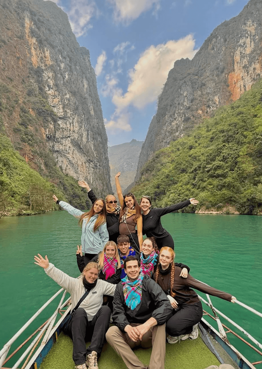 A small group of smiling tourists waving from the bow of a boat as they cruise down the bright emerald waters of the Nho Que River, surrounded by towering, steep karst cliffs in Ha Giang.