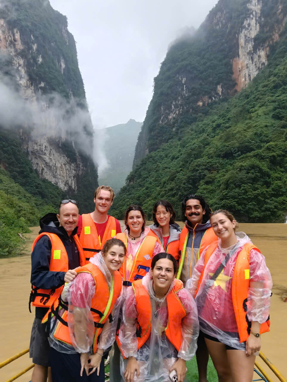 A smiling group of tourists wearing orange life jackets and rain ponchos taking a boat ride through the towering, misty Tu San Alley canyon on the Nho Que River in Ha Giang.