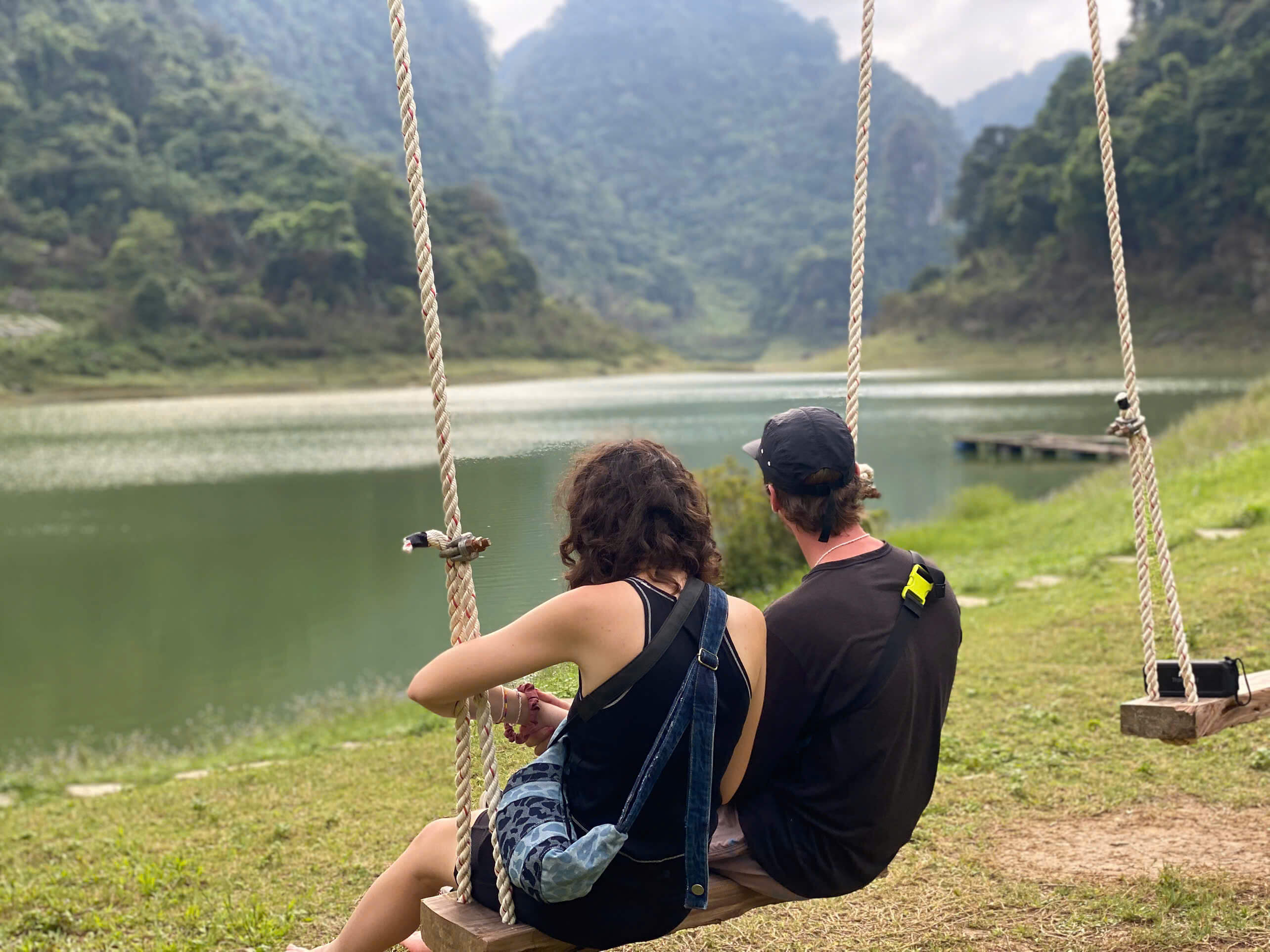 A couple sitting closely together on a rustic wooden swing, facing a completely serene, emerald green lake surrounded by quiet mountains and lush forests on the Cao Bang Loop.