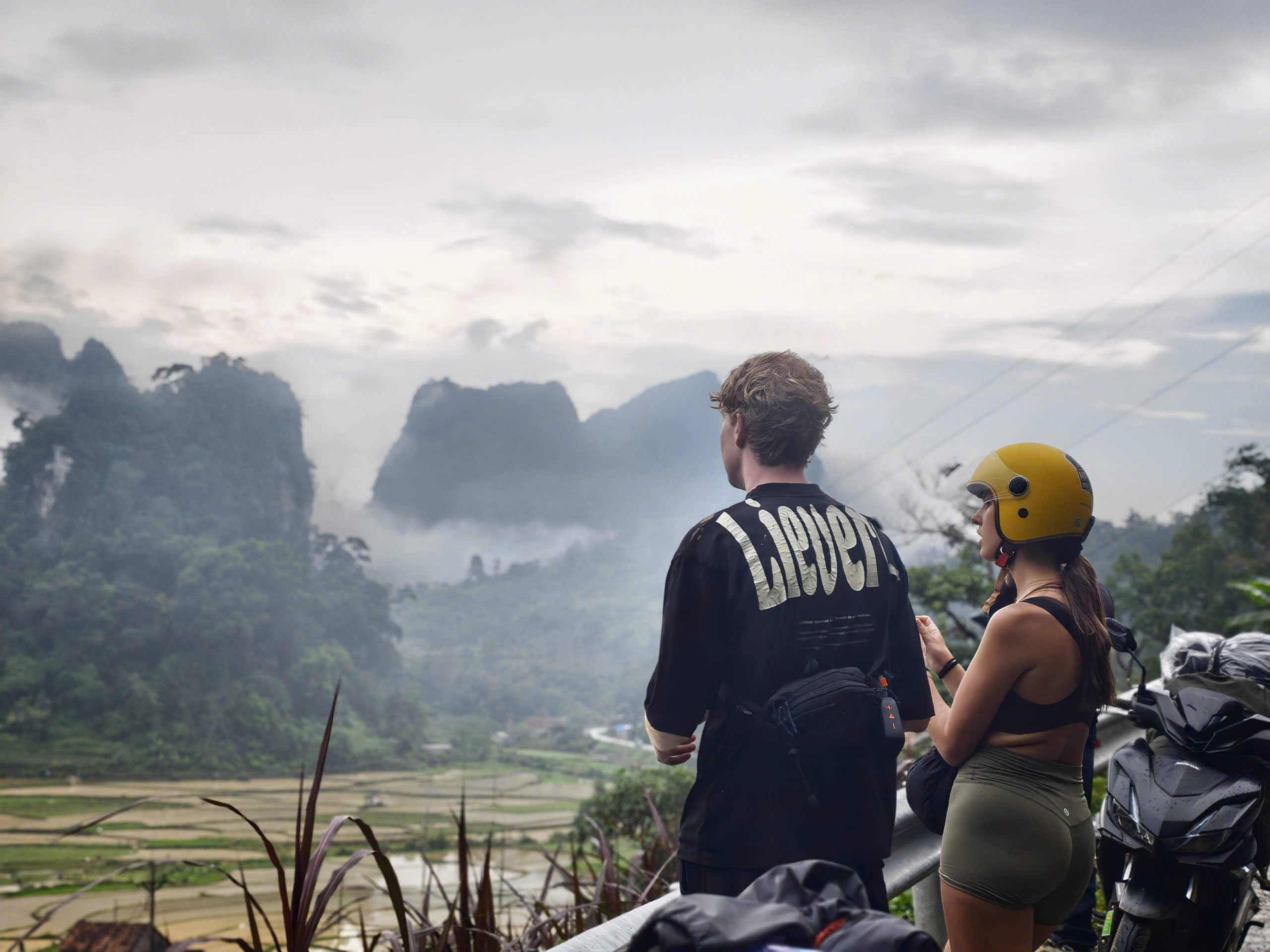 A couple taking a break from their motorbike ride, looking out over a breathtaking, misty valley filled with rice paddies and towering limestone karst mountains in Northern Vietnam.