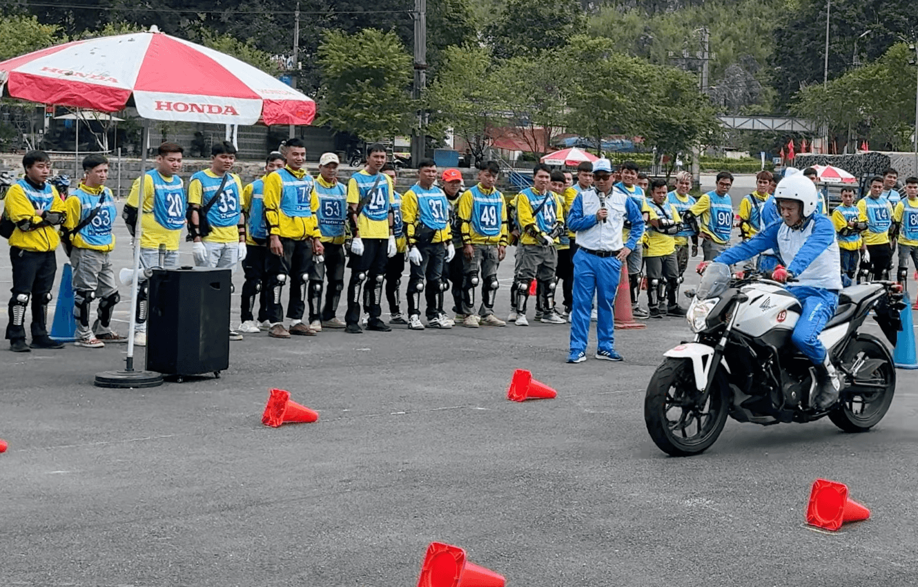 A line of Motorventures Easy Riders wearing yellow uniforms and protective knee pads watch closely as a Honda Vietnam master instructor demonstrates advanced maneuvering techniques on a cone-marked obstacle course.