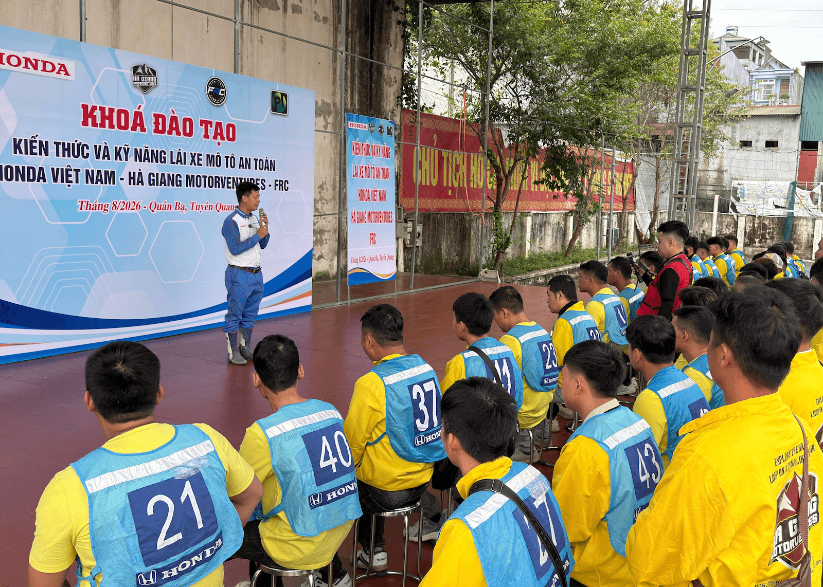 A Honda Vietnam expert with a microphone leads a classroom theory session for a large group of seated Ha Giang Motorventures guides in front of an official training banner.