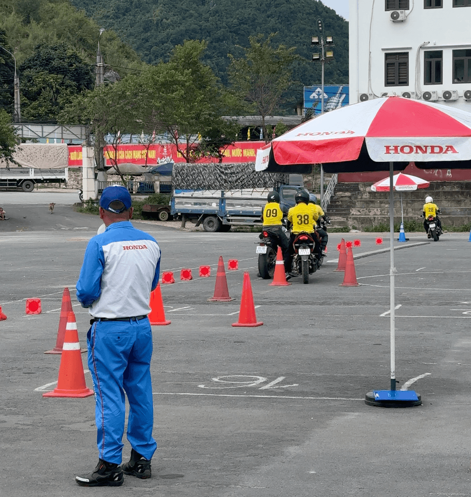 A Honda Vietnam instructor in a blue uniform observes two Ha Giang Motorventures Easy Riders practicing safe pillion passenger riding techniques on a closed obstacle course marked by red traffic cones.